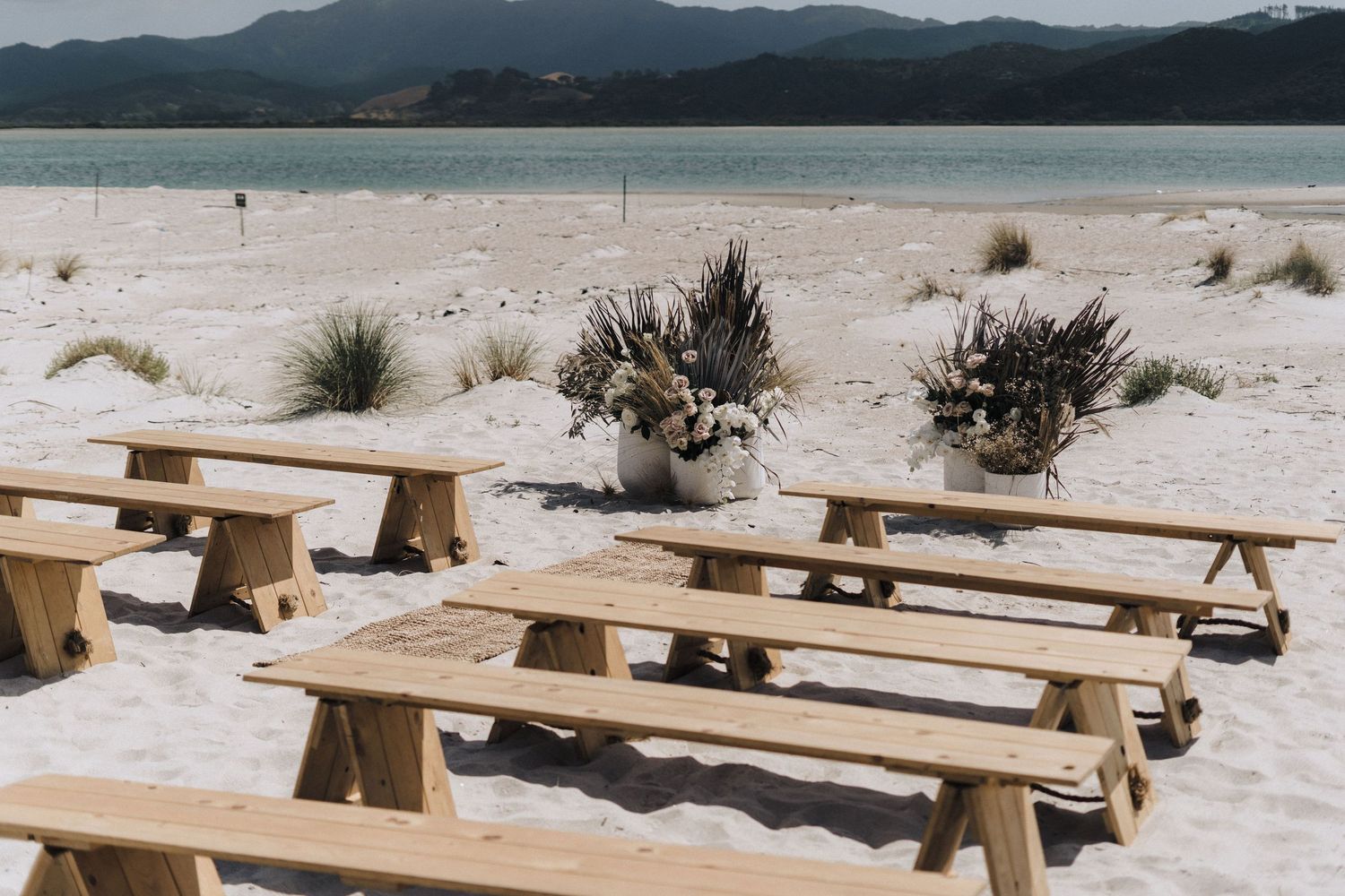 Wooden picnic tables arranged on a white sandy beach with mountains and turquoise water in the background.