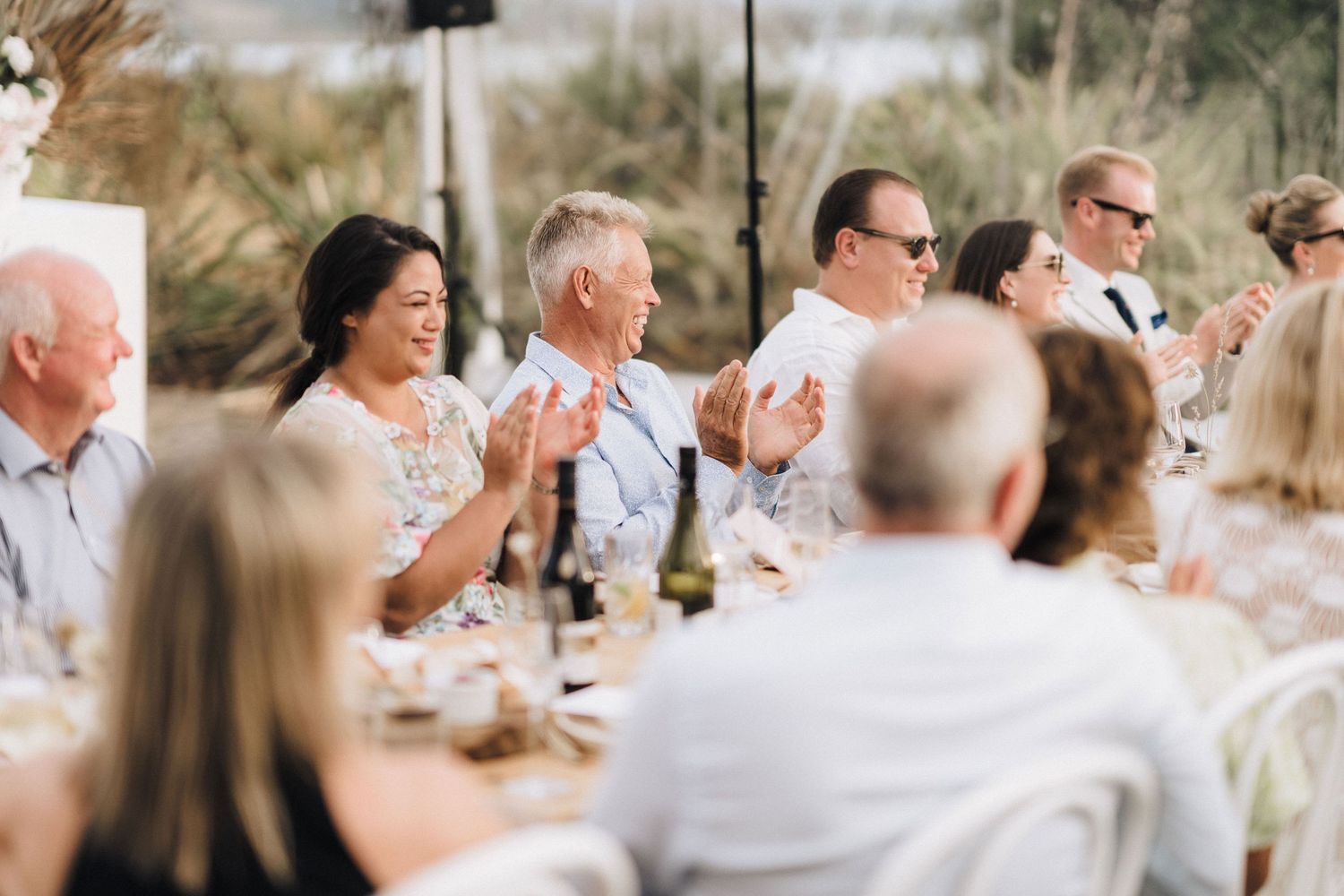 Wedding guests seated at an outdoor reception table applaud enthusiastically during a celebration.
