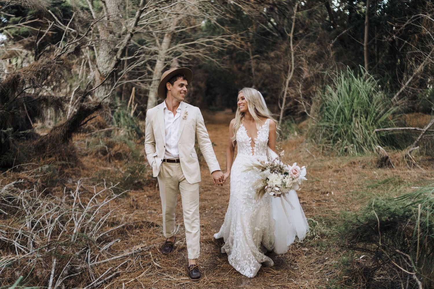 A couple walks hand in hand through a rustic outdoor setting wearing elegant wedding attire.