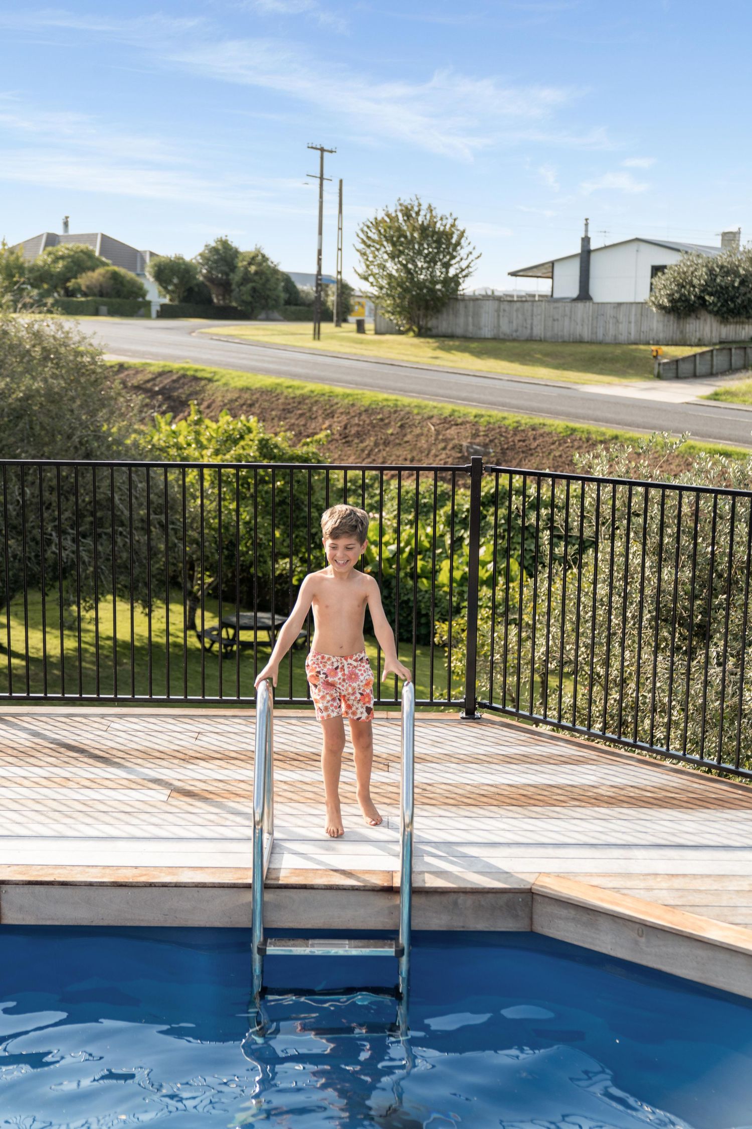 A summer scene shows someone standing at the edge of a bright blue swimming pool with metal railings.