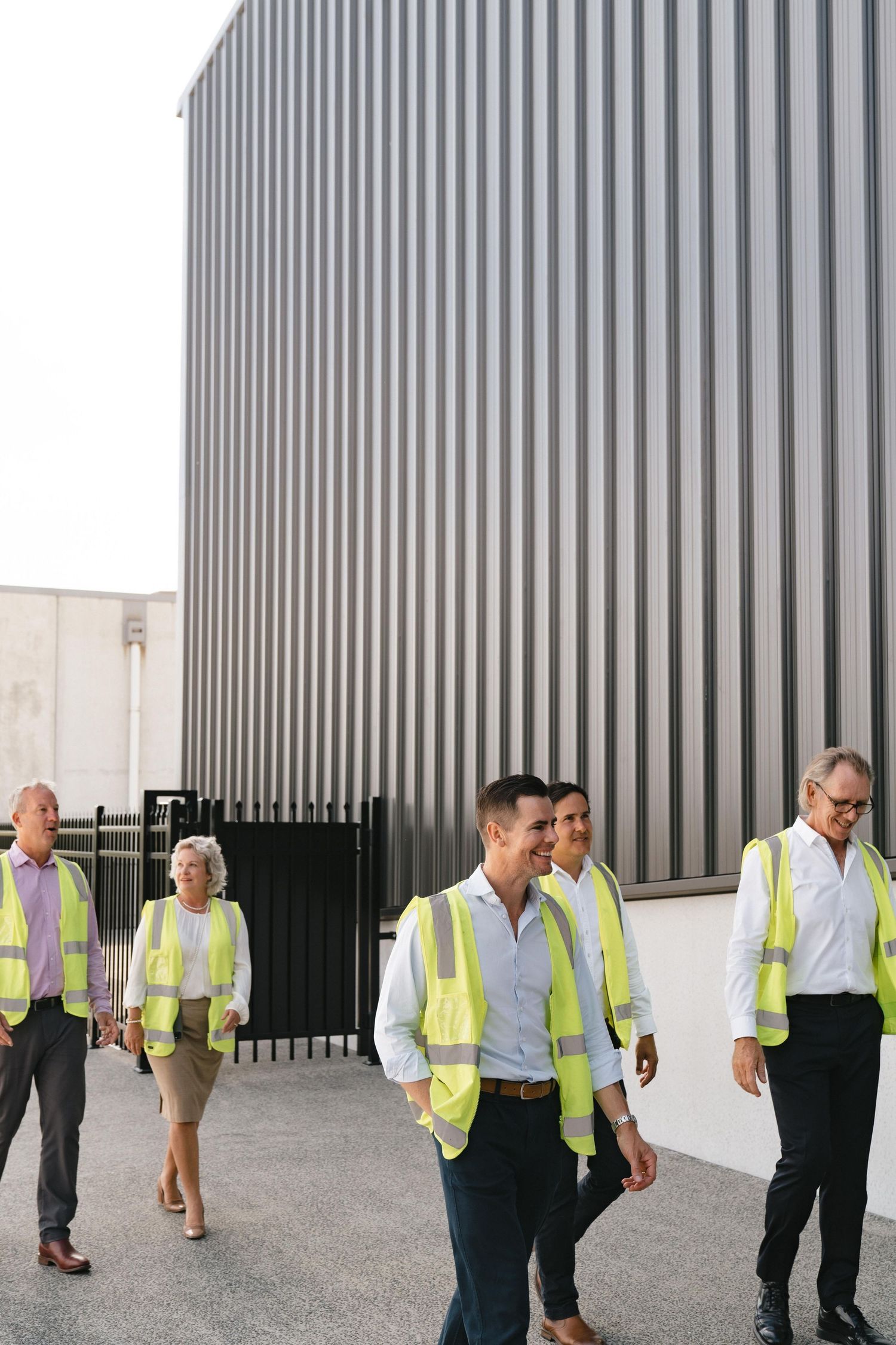Workers in high visibility vests inspect a modern building with large steel columns during a construction site walkthrough.