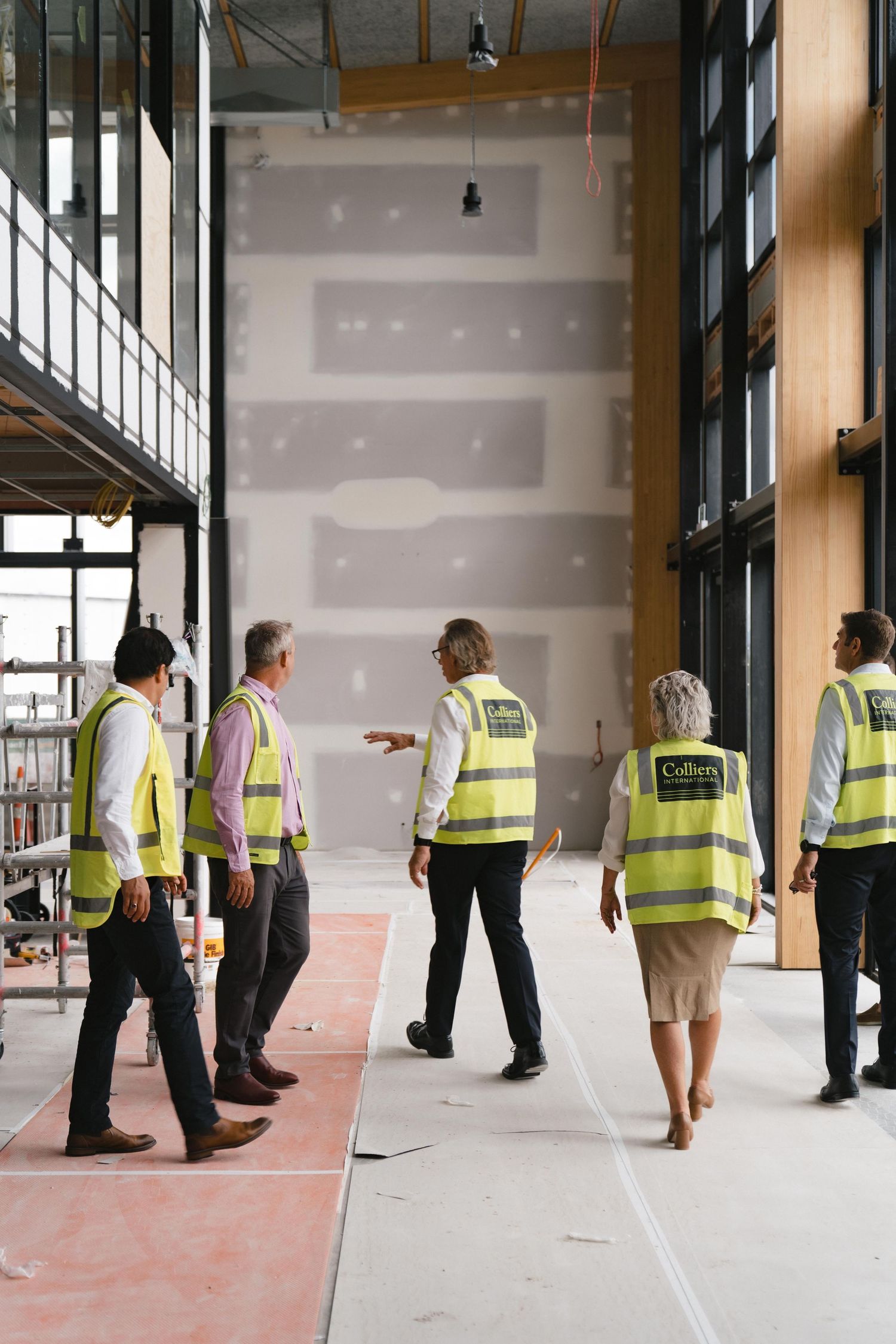 Construction workers wearing reflective safety vests examine interior space with concrete floors and exposed walls.