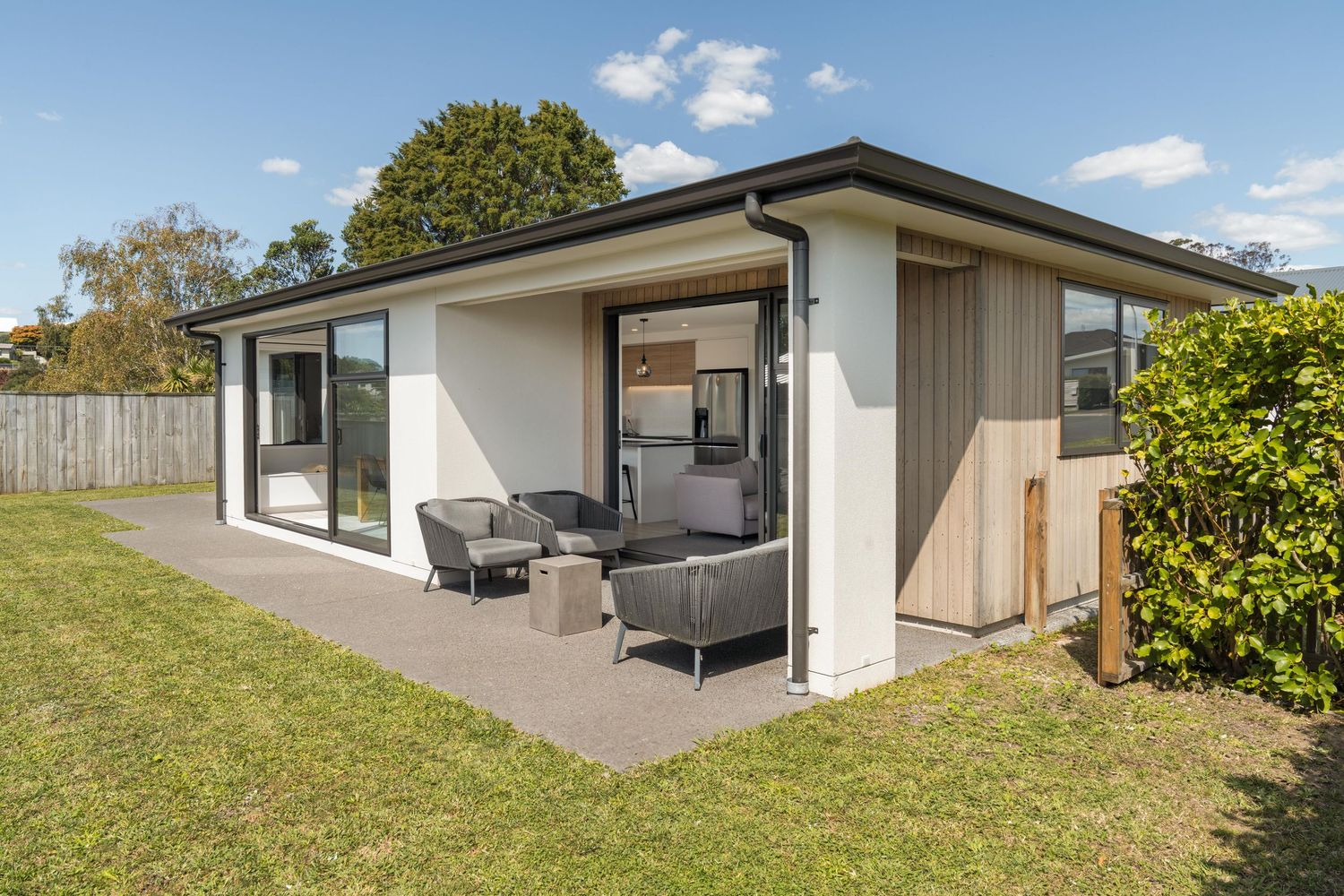 Modern white house exterior with covered patio area and outdoor furniture on concrete patio surrounded by green lawn.