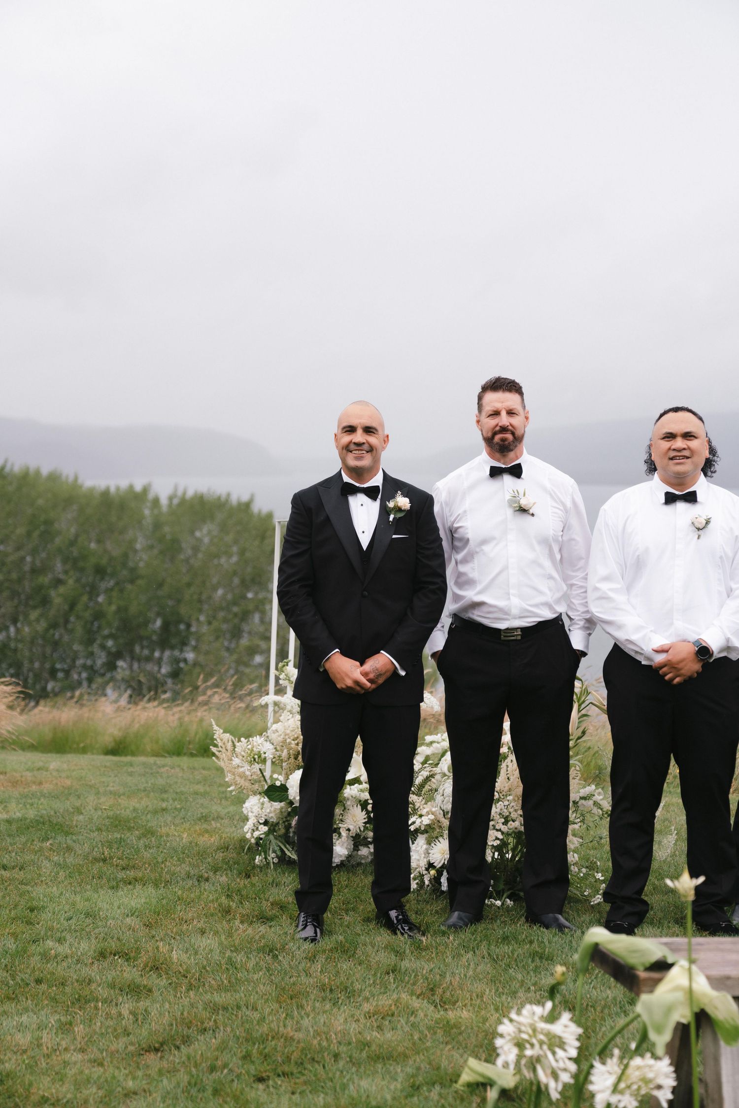 Groomsmen in black tuxedos and bowties pose together on a grassy hilltop during an outdoor wedding.