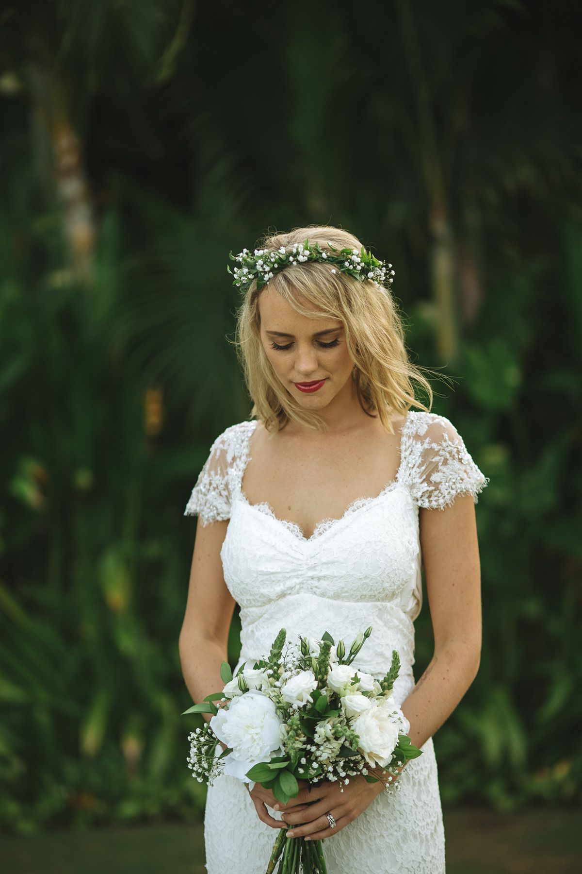 A bride in lace cap sleeve dress holds white bouquet in lush garden setting.