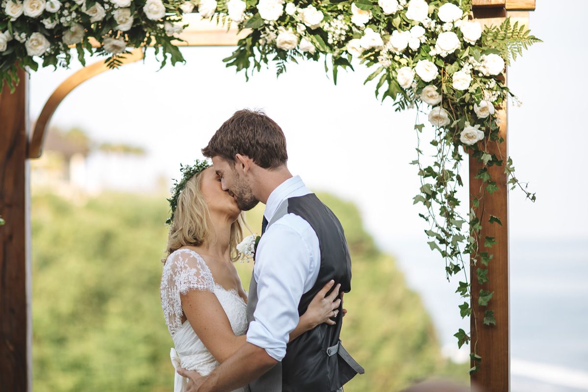 A romantic moment under a floral arch with white flowers and greenery during an outdoor wedding ceremony.
