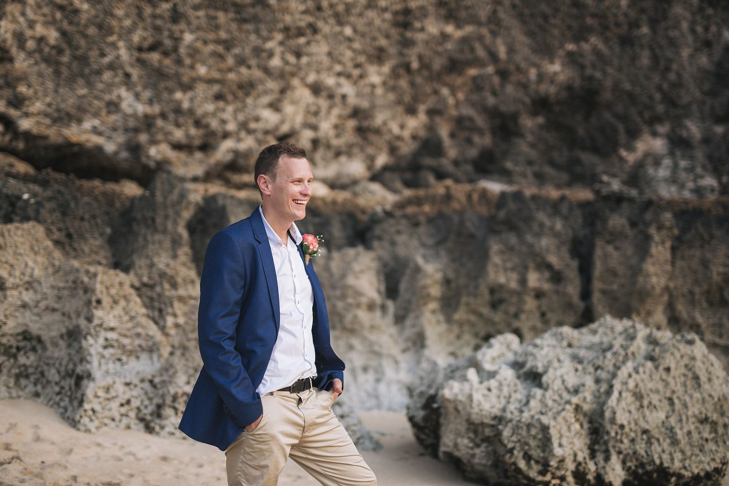 Groom in navy blazer and khaki pants standing against rocky coastal backdrop.