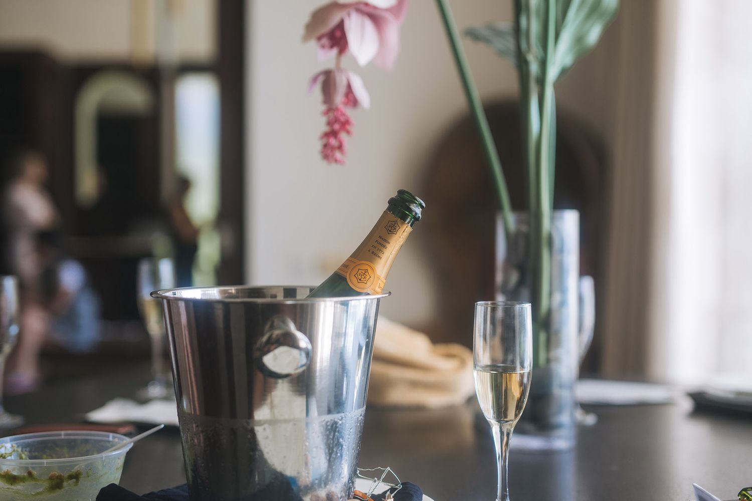 Champagne bottle chilling in silver ice bucket with flutes and floral decorations on elegant table setting.