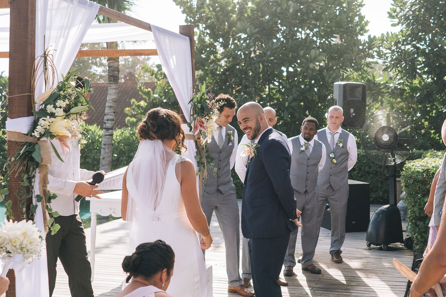 Wedding ceremony guests and wedding party gathered under an outdoor white canopy on a sunny day.