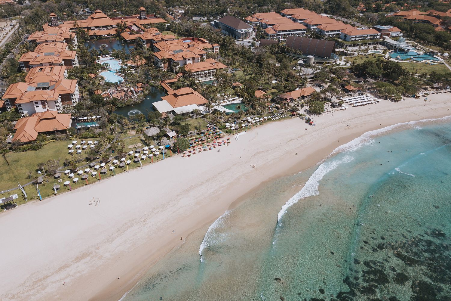 Aerial view of luxury beachfront resort with terracotta roofs along turquoise waters and white sandy beach.