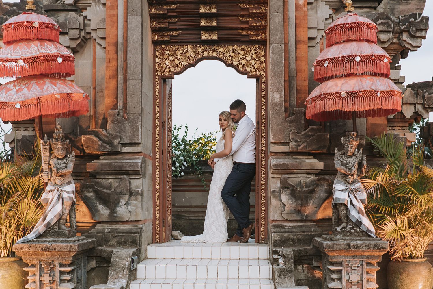 Traditional Balinese temple ceremony with red umbrellas and ornate stone architecture at sunset.