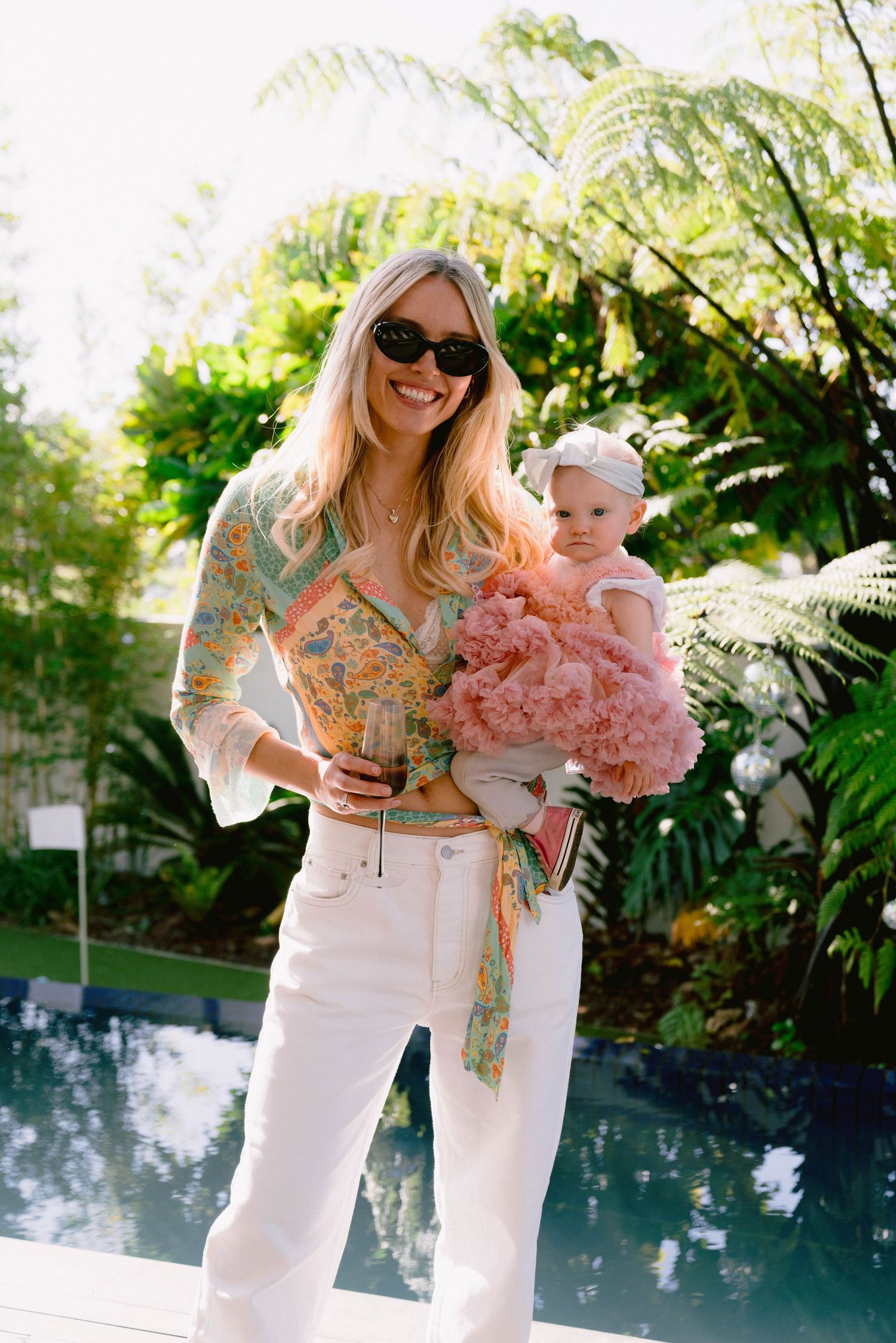 Woman in floral top and white pants holds baby in pink ruffled dress by tropical poolside.