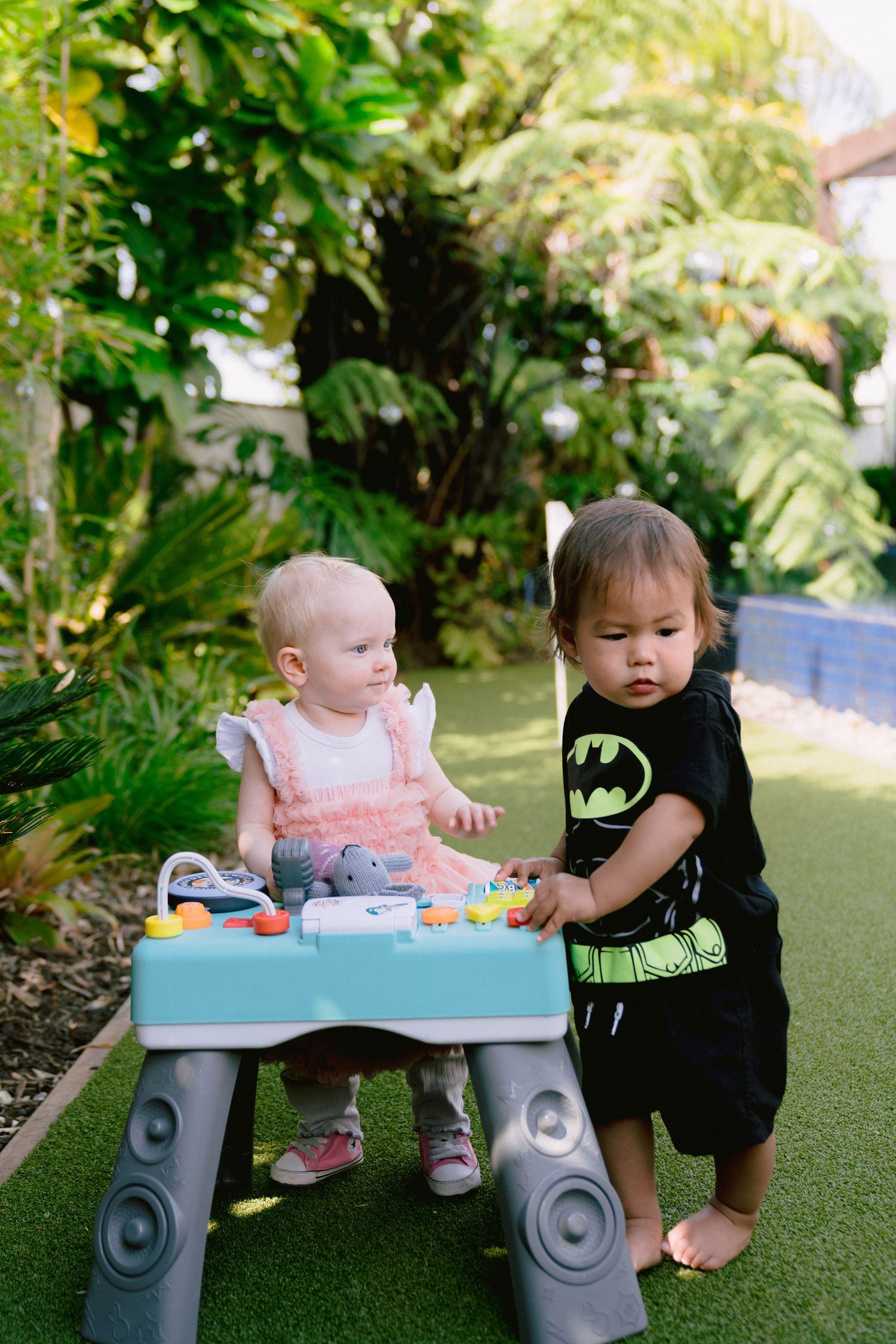 Two toddlers play at a blue activity table on artificial grass surrounded by lush tropical plants.