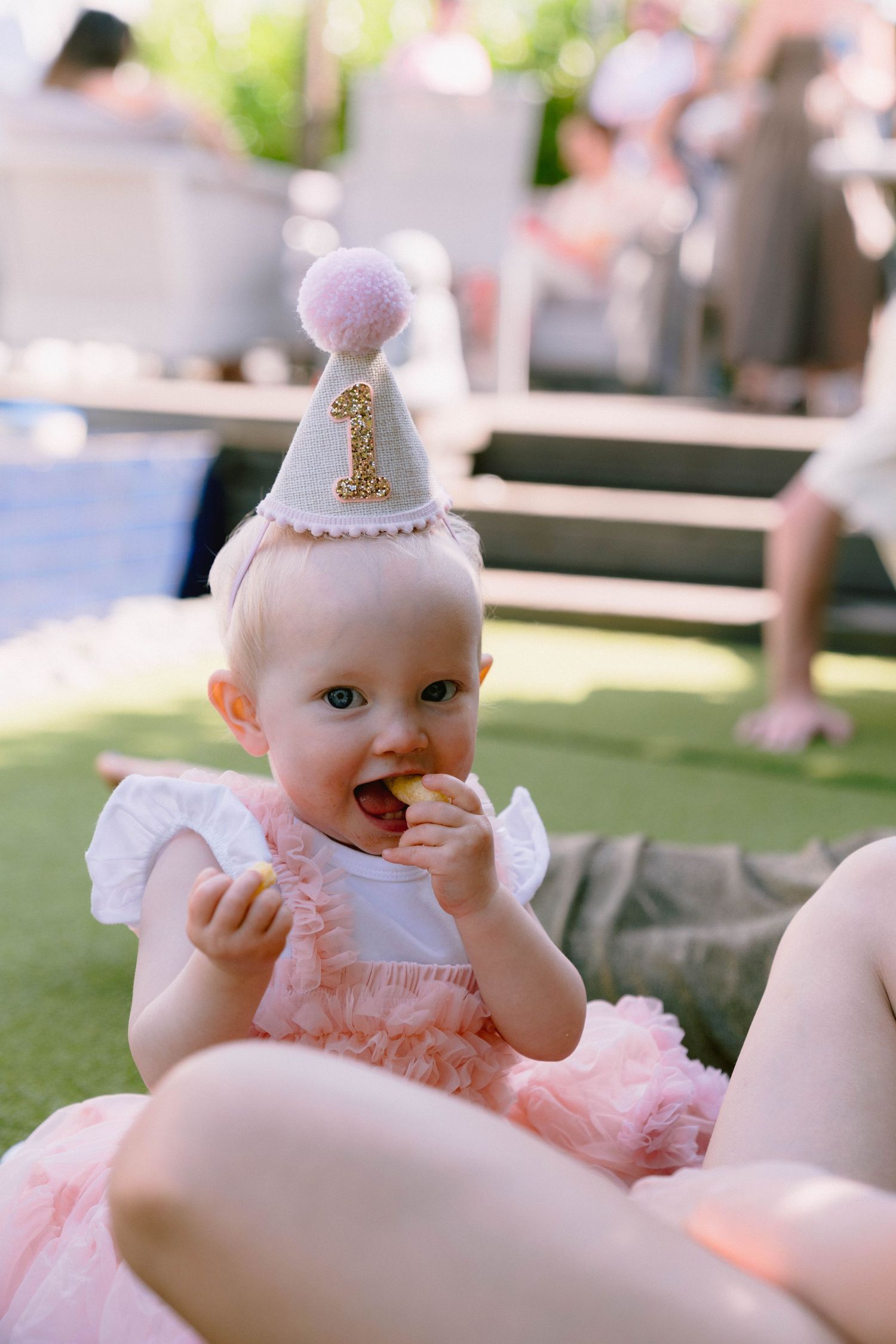 A baby wearing a pink tulle dress and small crown sits on a white surface during a special celebration.