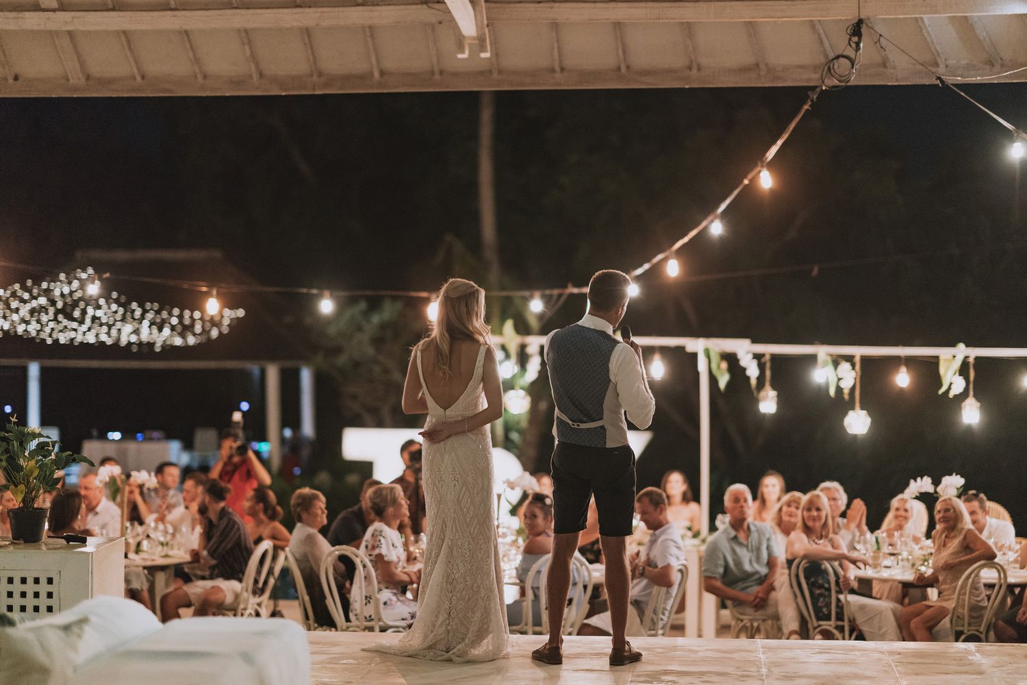Newlyweds address their wedding guests under romantic string lights at an outdoor reception.