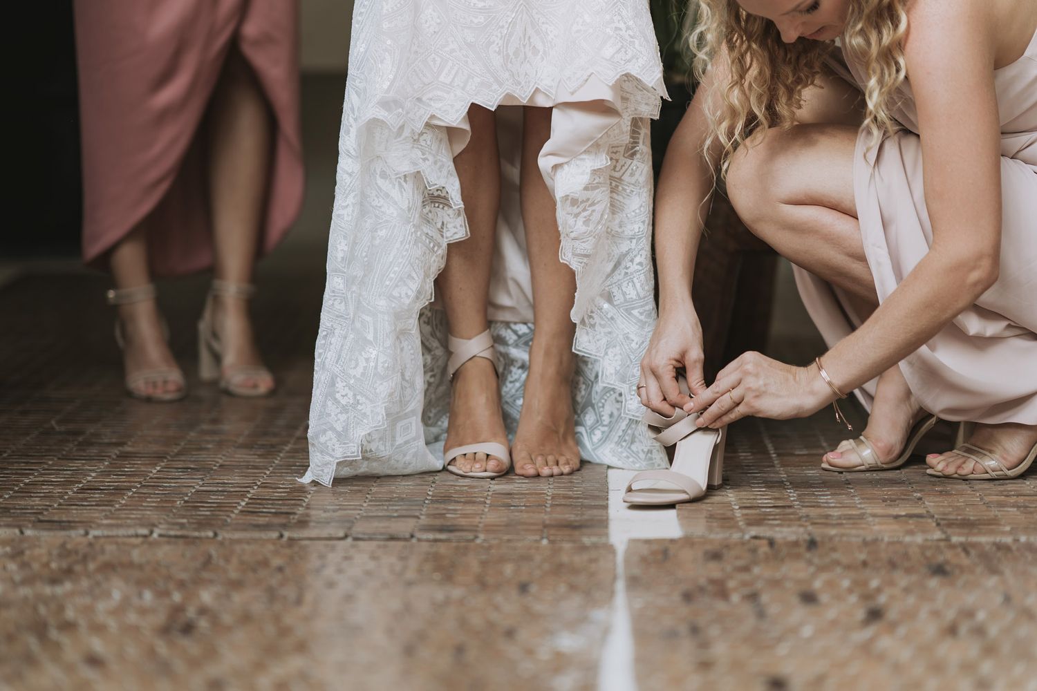 Wedding bridesmaids in light dresses showing off their shoes in a casual row against a rustic wall background.