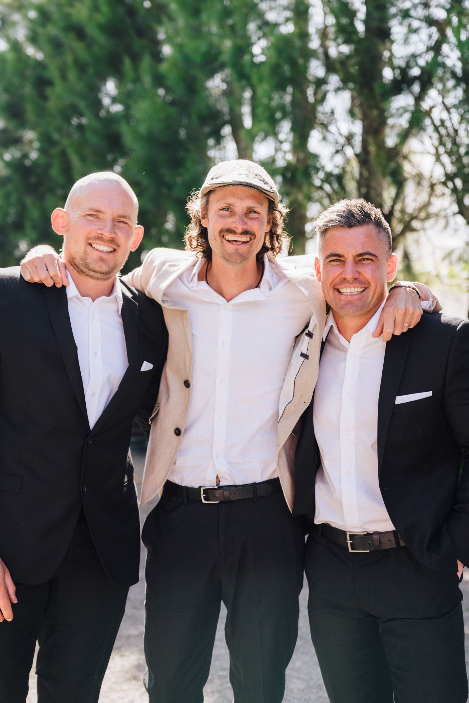 Three men standing together outdoors smiling, two in dark suits and one in light colored attire.