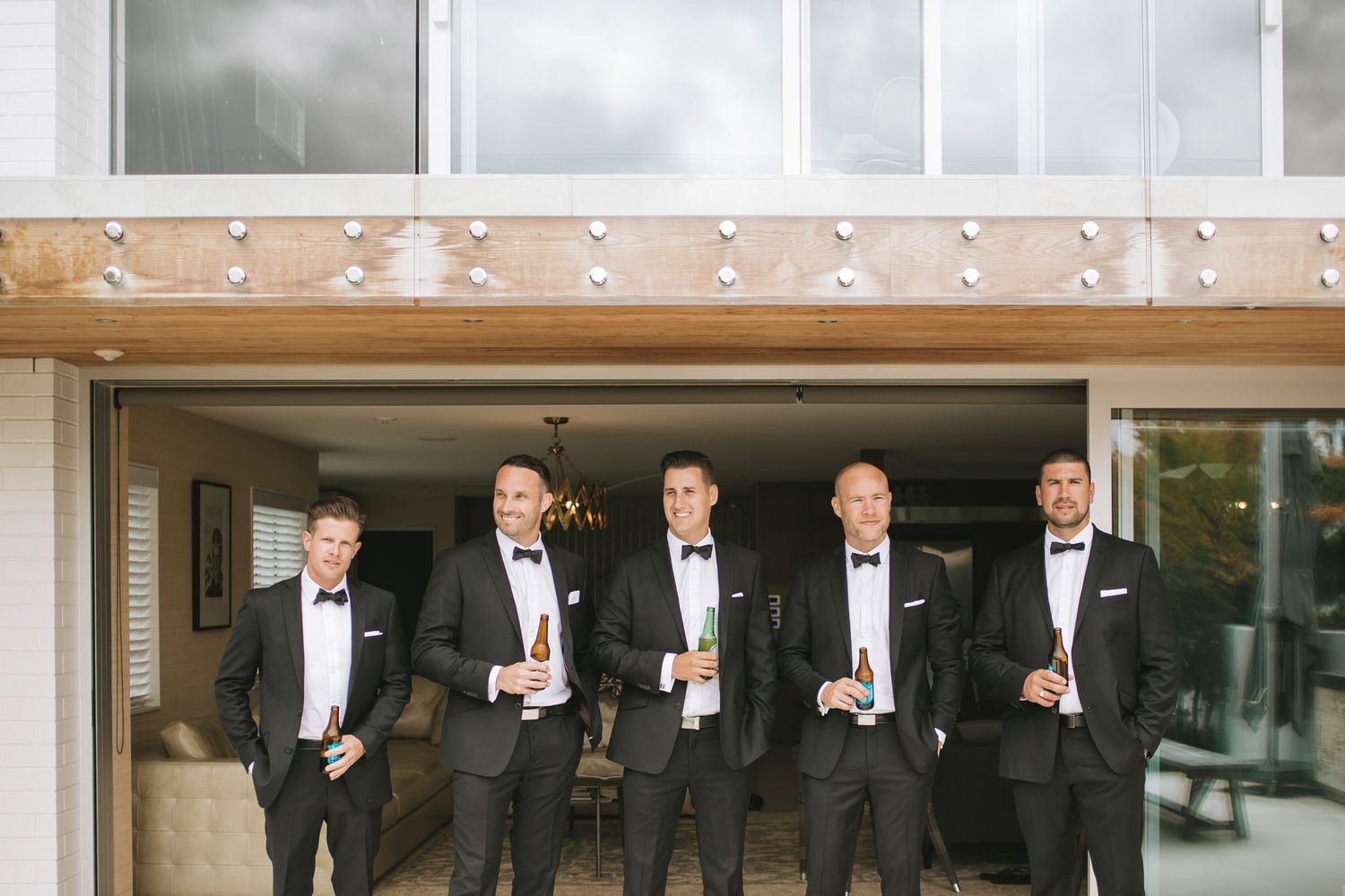 Groomsmen in black tuxedos and bowties stand together holding drinks outside a building entrance.