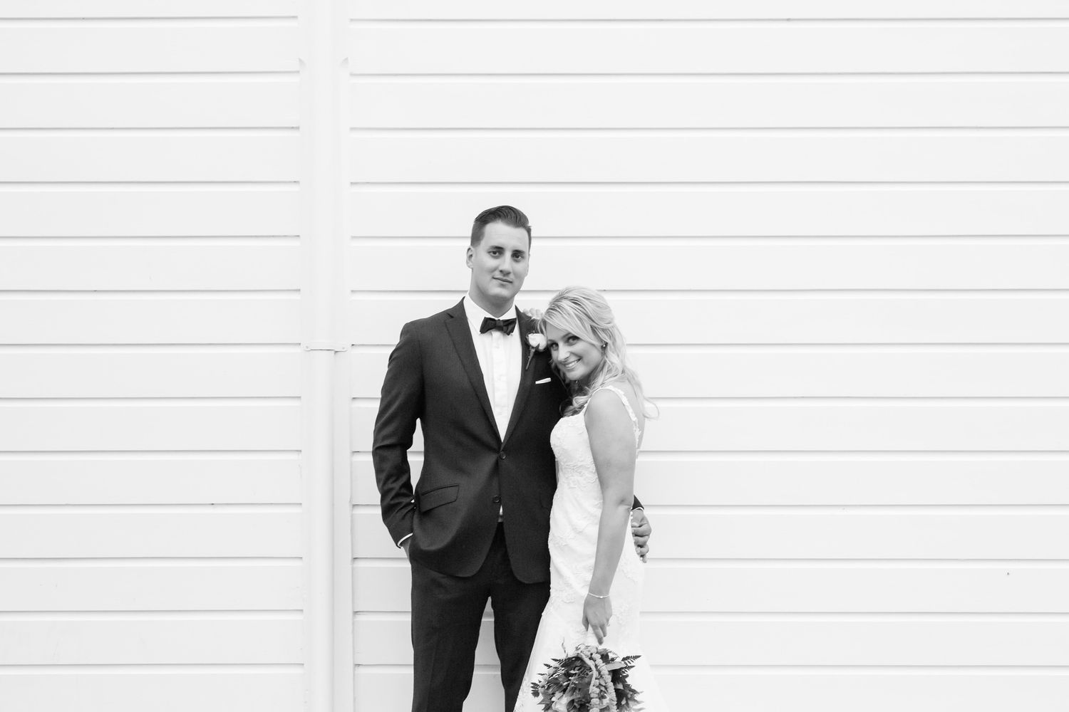 Wedding couple poses against white wall in elegant black and white portrait with bride holding bouquet.