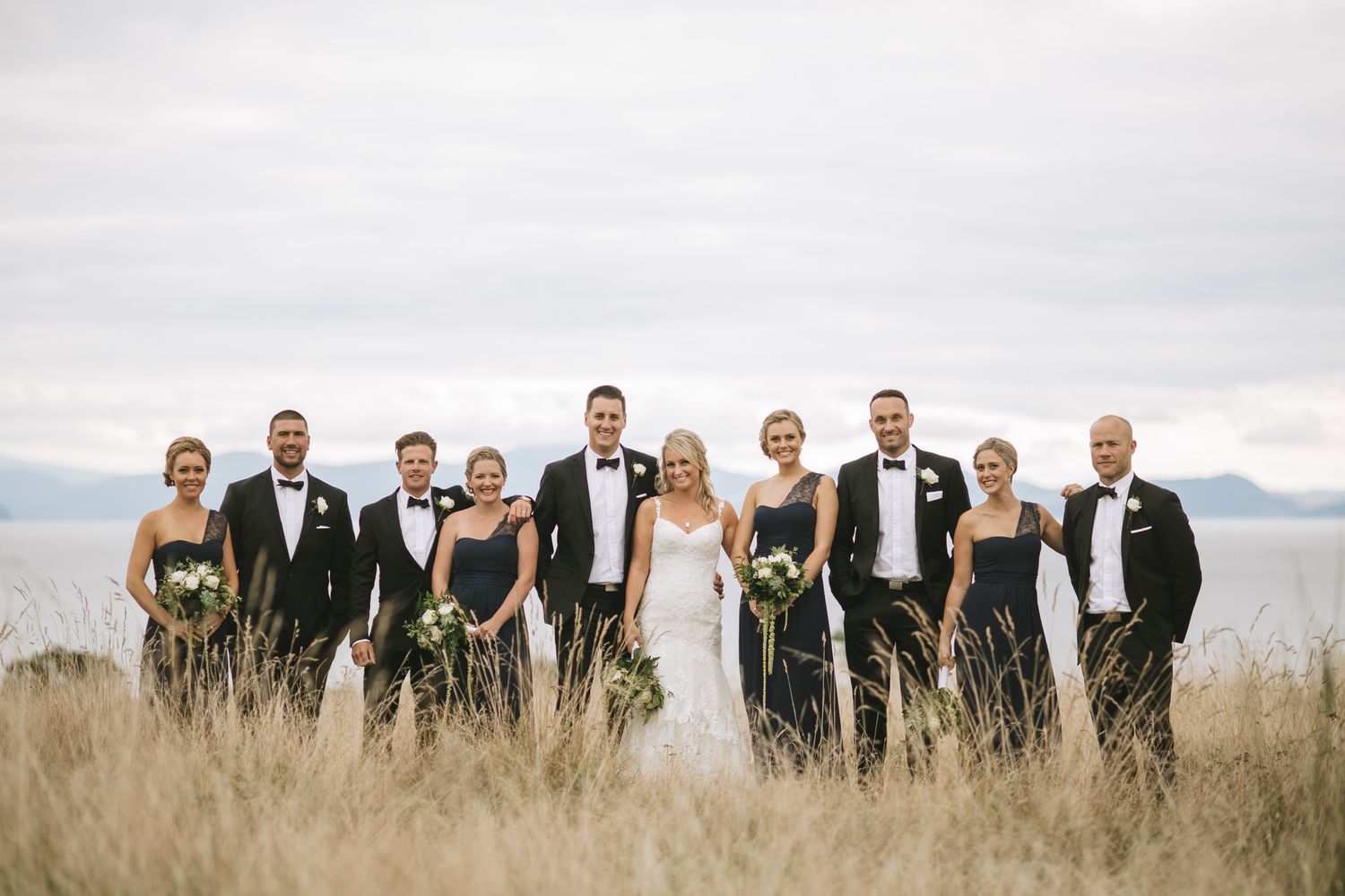 Wedding party in formal attire standing together in tall grass field with mountain landscape in background.