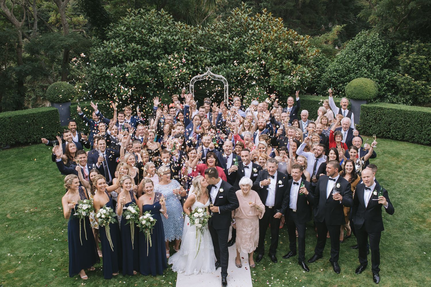 Large wedding party gathers on a green lawn surrounded by lush gardens for formal group photograph.