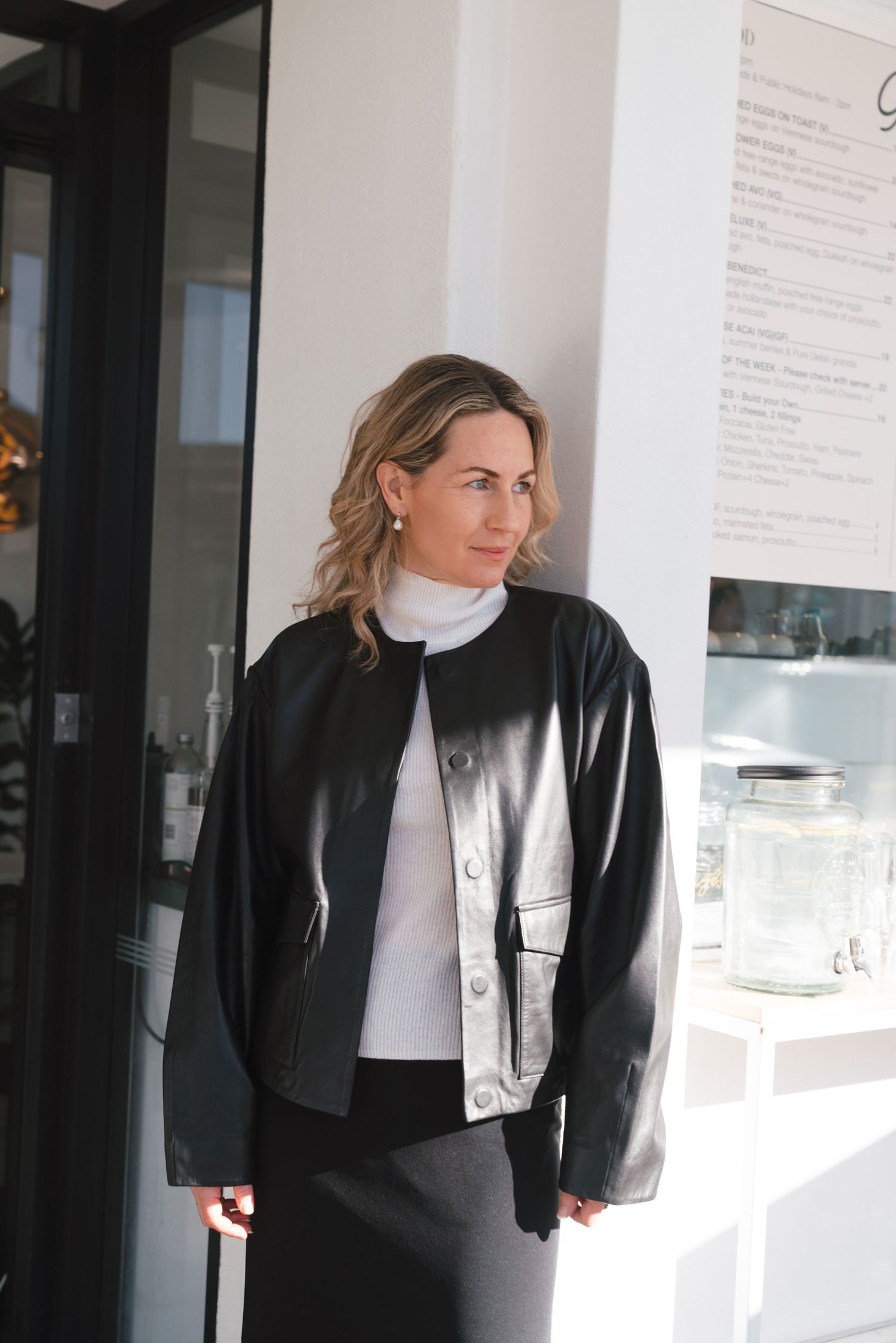 Fashion model wearing a black leather jacket over white turtleneck sweater in a modern interior setting.