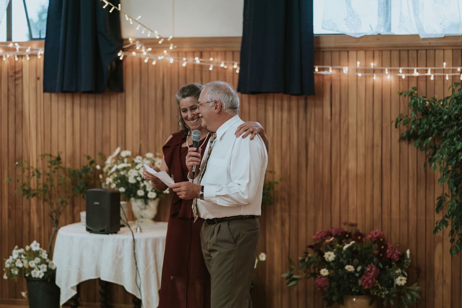 Two people share an emotional moment during a ceremony in a rustic venue decorated with fairy lights and floral arrangements.