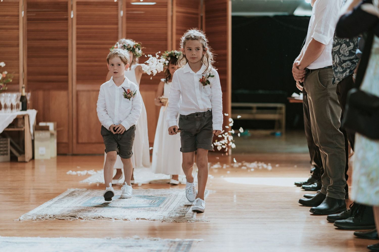 Two young attendants in matching white shirts and shorts skip down the wedding aisle runner at an indoor ceremony.