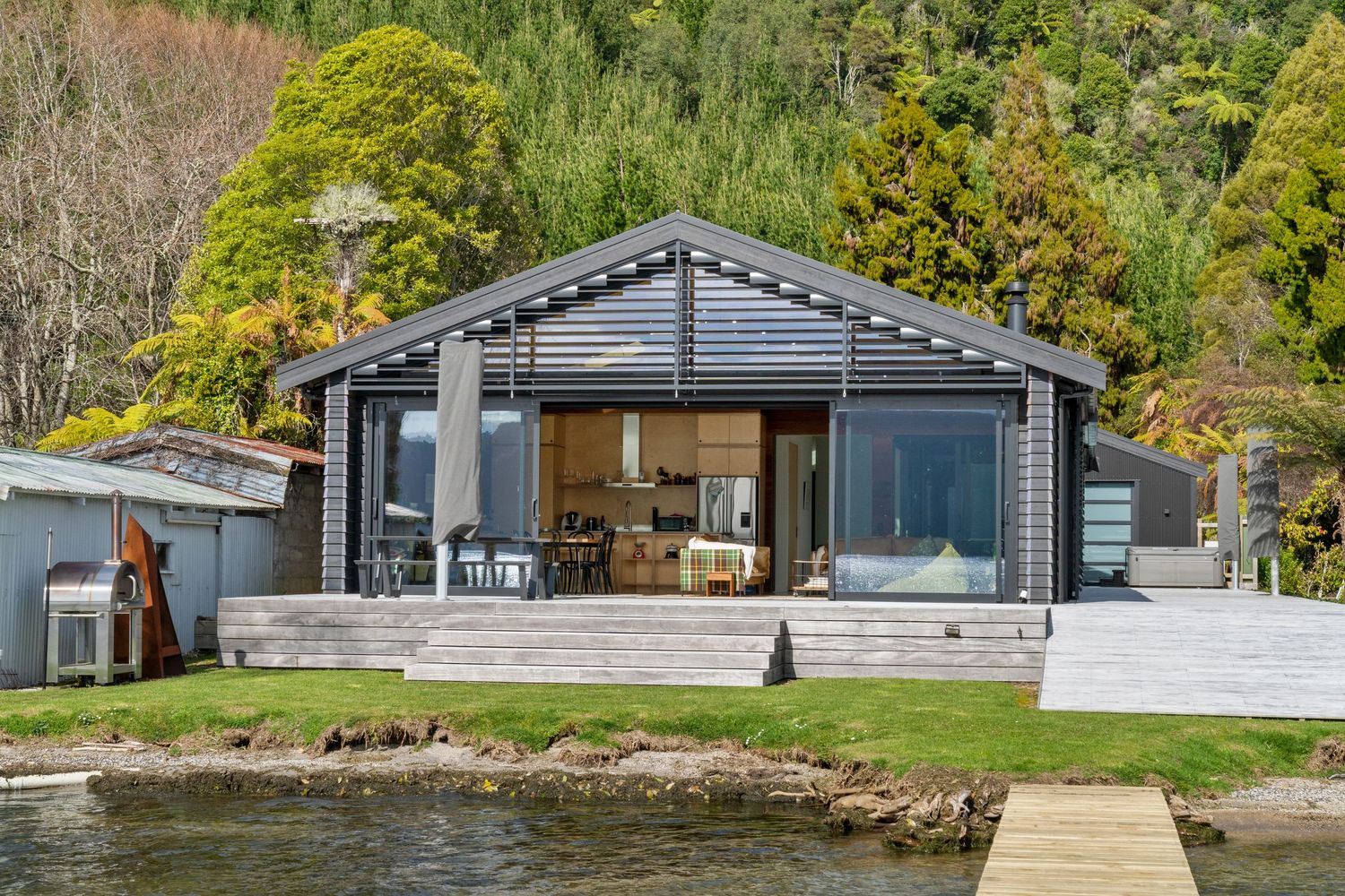 Modern lakeside cabin with gray exterior, large glass doors, and spacious deck overlooking calm waters.