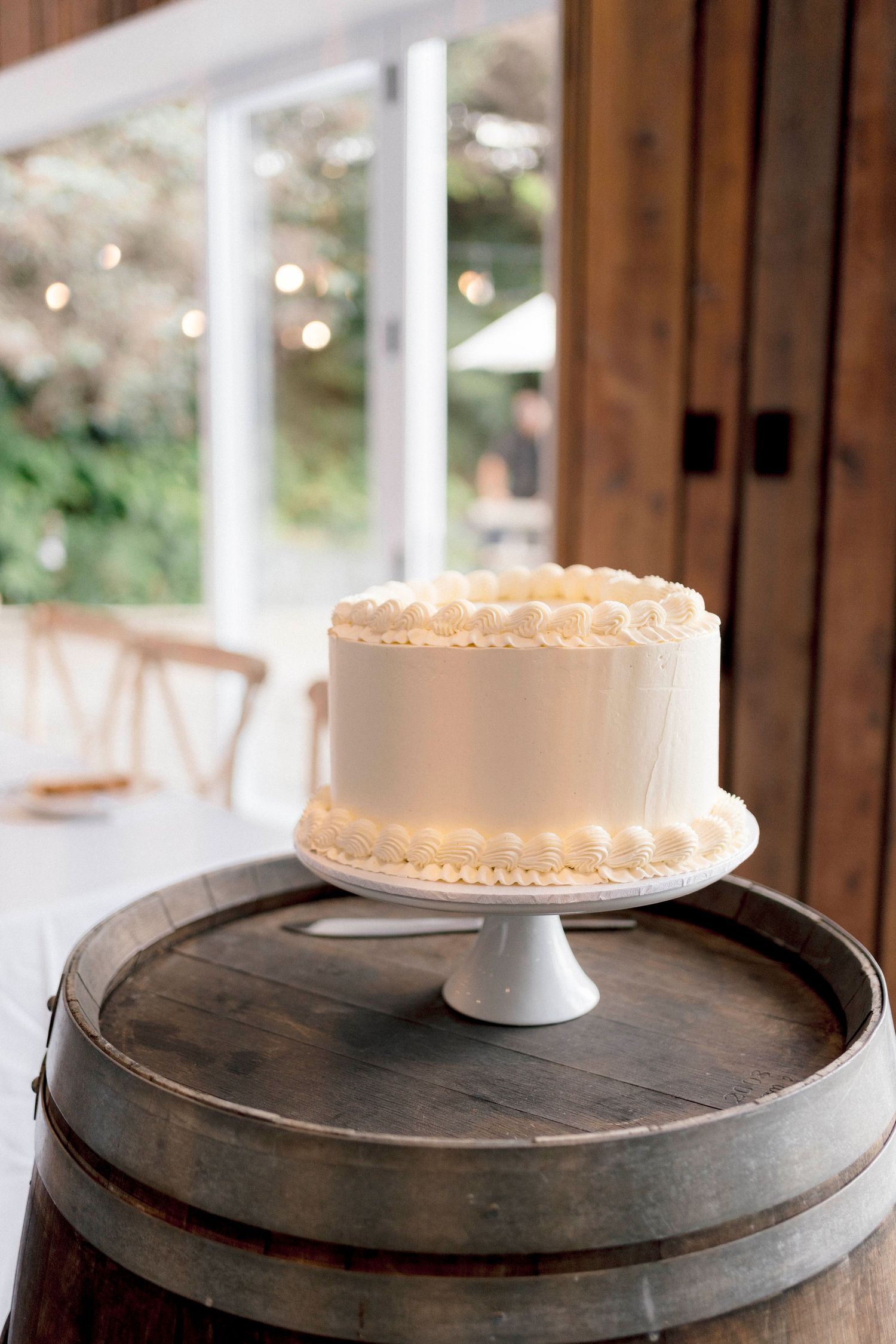 A simple white wedding cake with piped details sits atop a wooden barrel display.