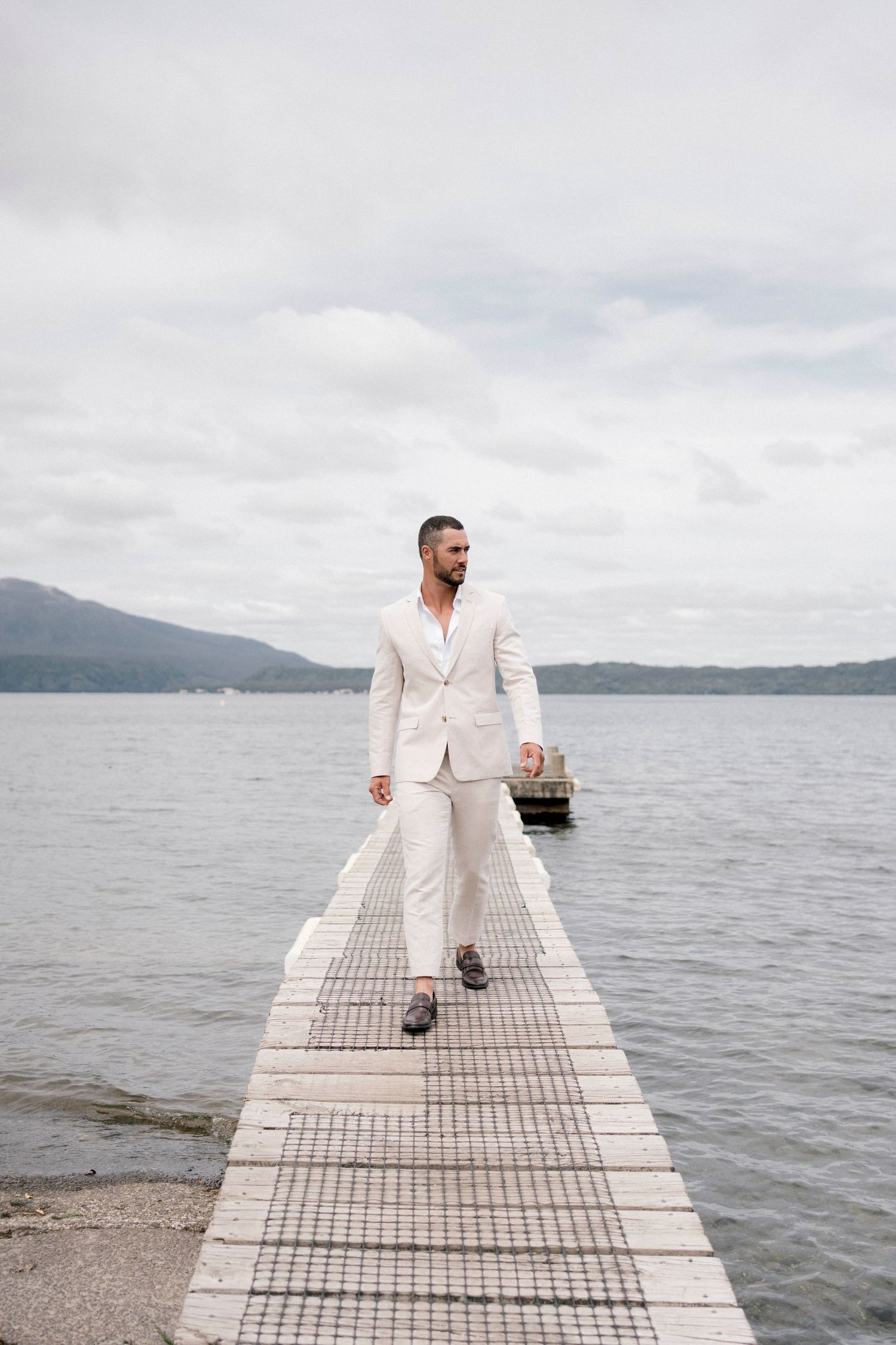 A person in white attire walking along a wooden dock over a serene lake with mountains in the background.