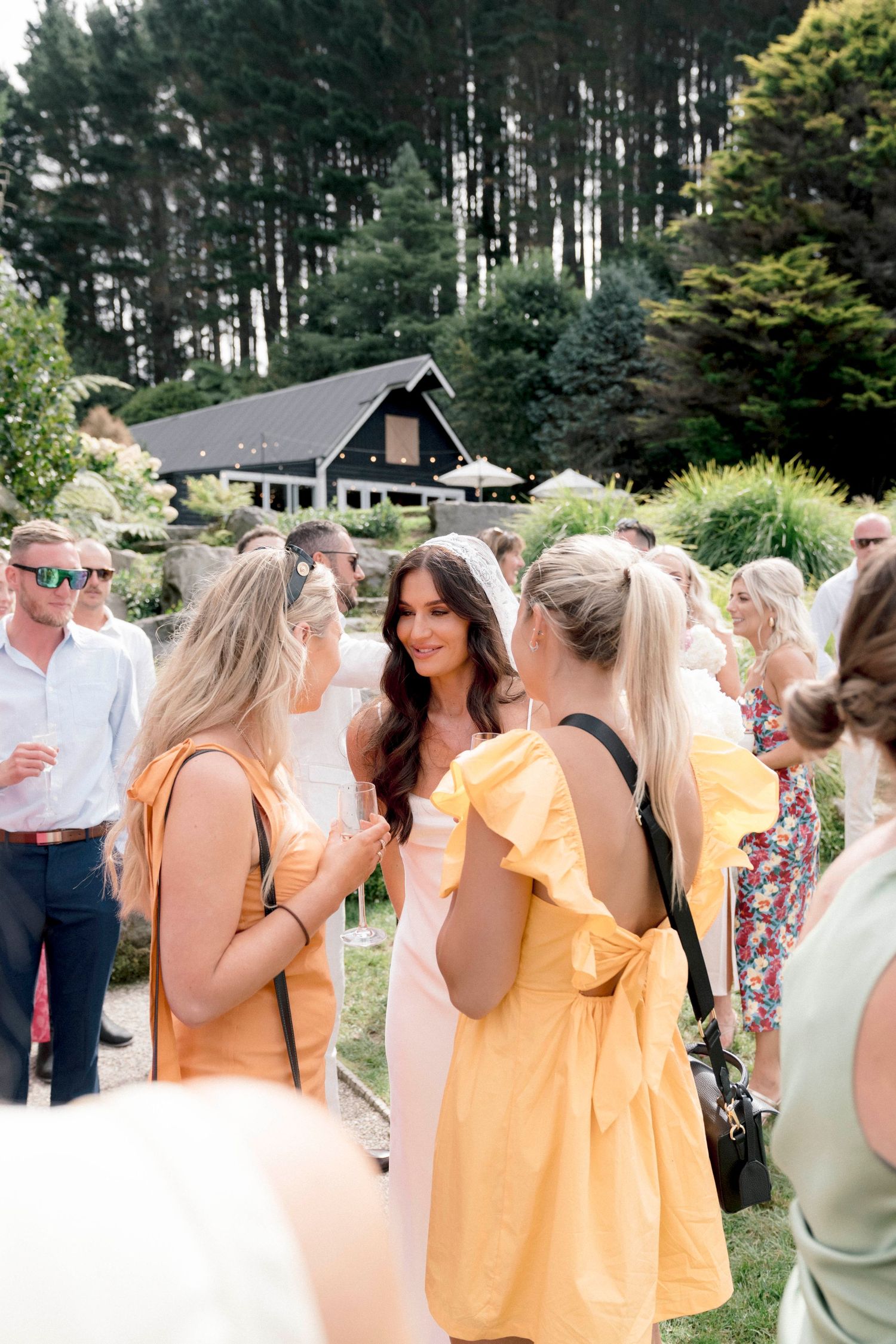 Guests mingle at an outdoor wedding reception with a rustic cabin backdrop surrounded by evergreen trees.