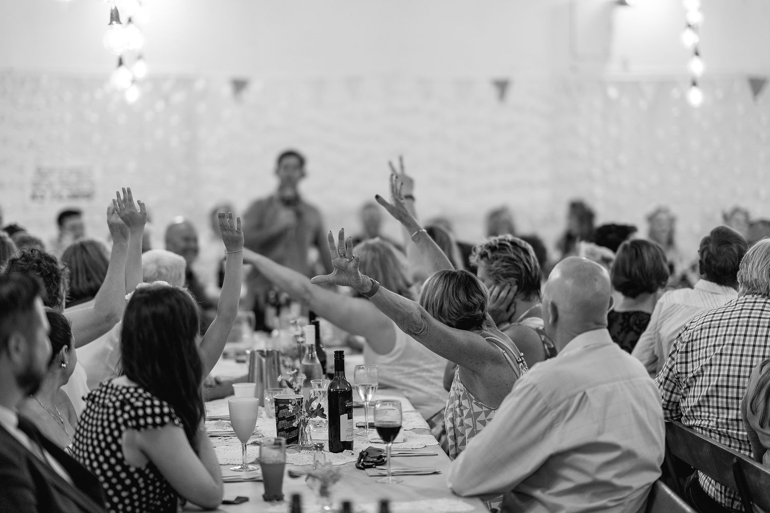 A black and white photo shows dinner guests raising their hands in celebration at a long banquet table.