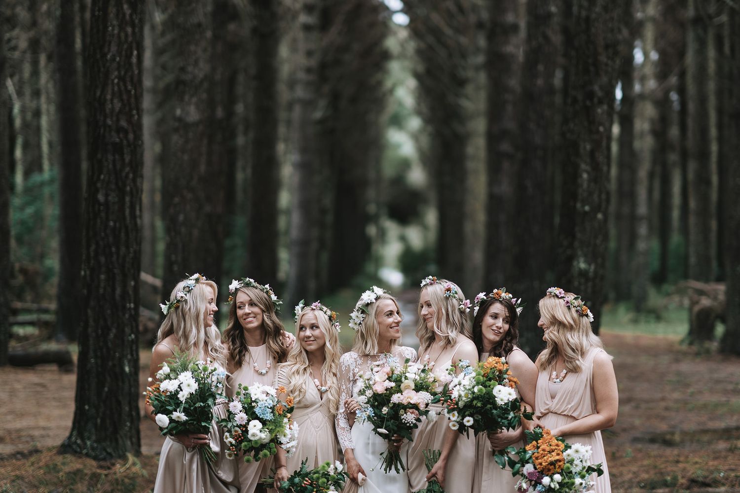 A group in matching beige dresses holds floral bouquets while standing between tall trees in a forest setting.