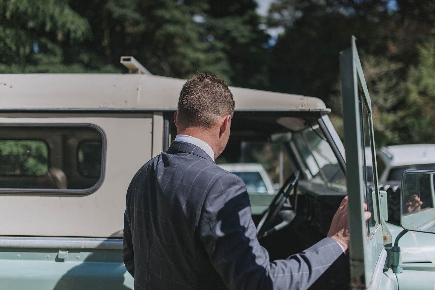 Groom in dark suit enters classic vintage car surrounded by evergreen trees on wedding day.