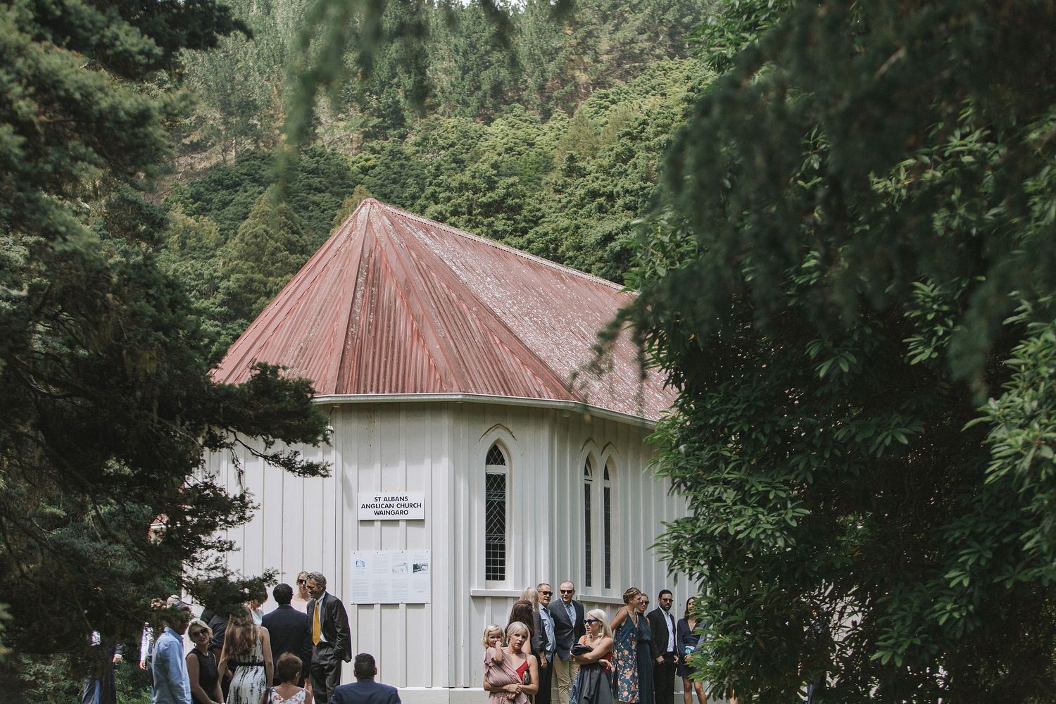 A row of small white rustic church buildings with brown roofs nestled among lush green trees.