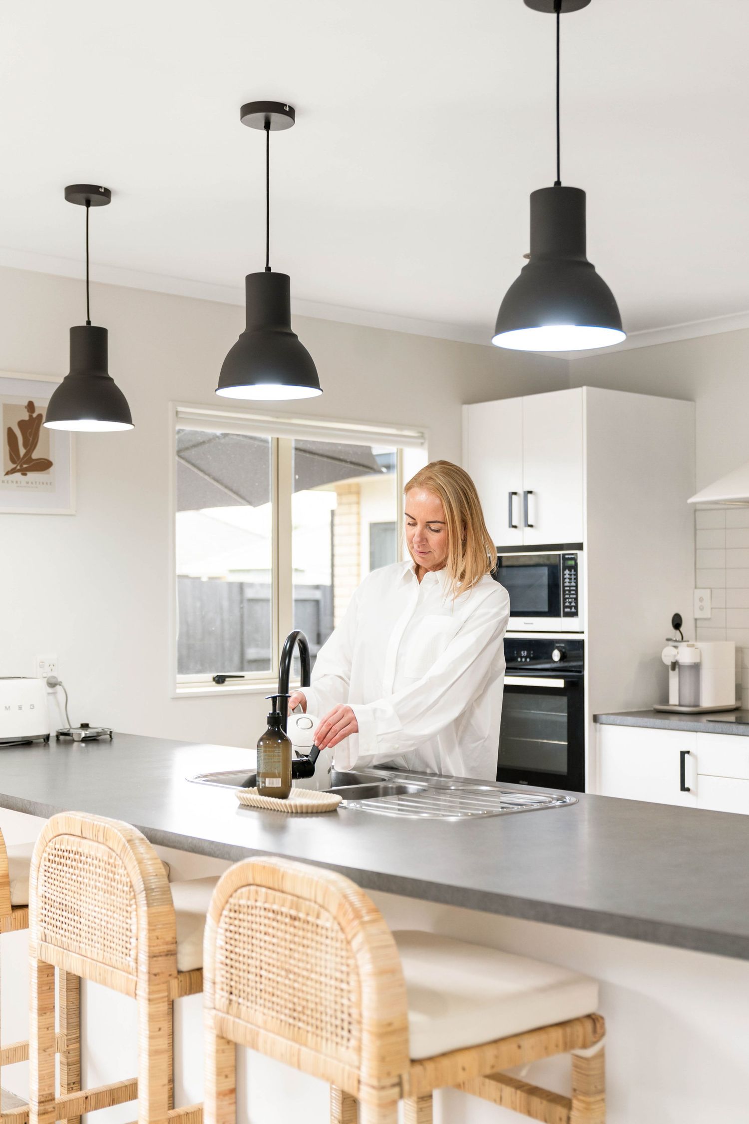 Modern white kitchen with black pendant lights and rattan bar stools at concrete countertop island.
