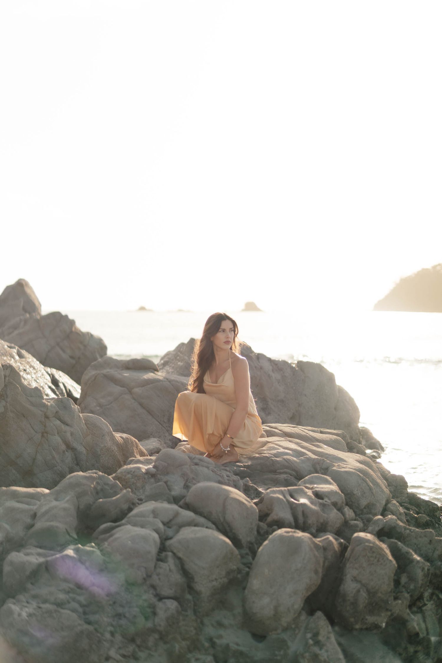 Person in light dress reclines on coastal rocks during golden hour.