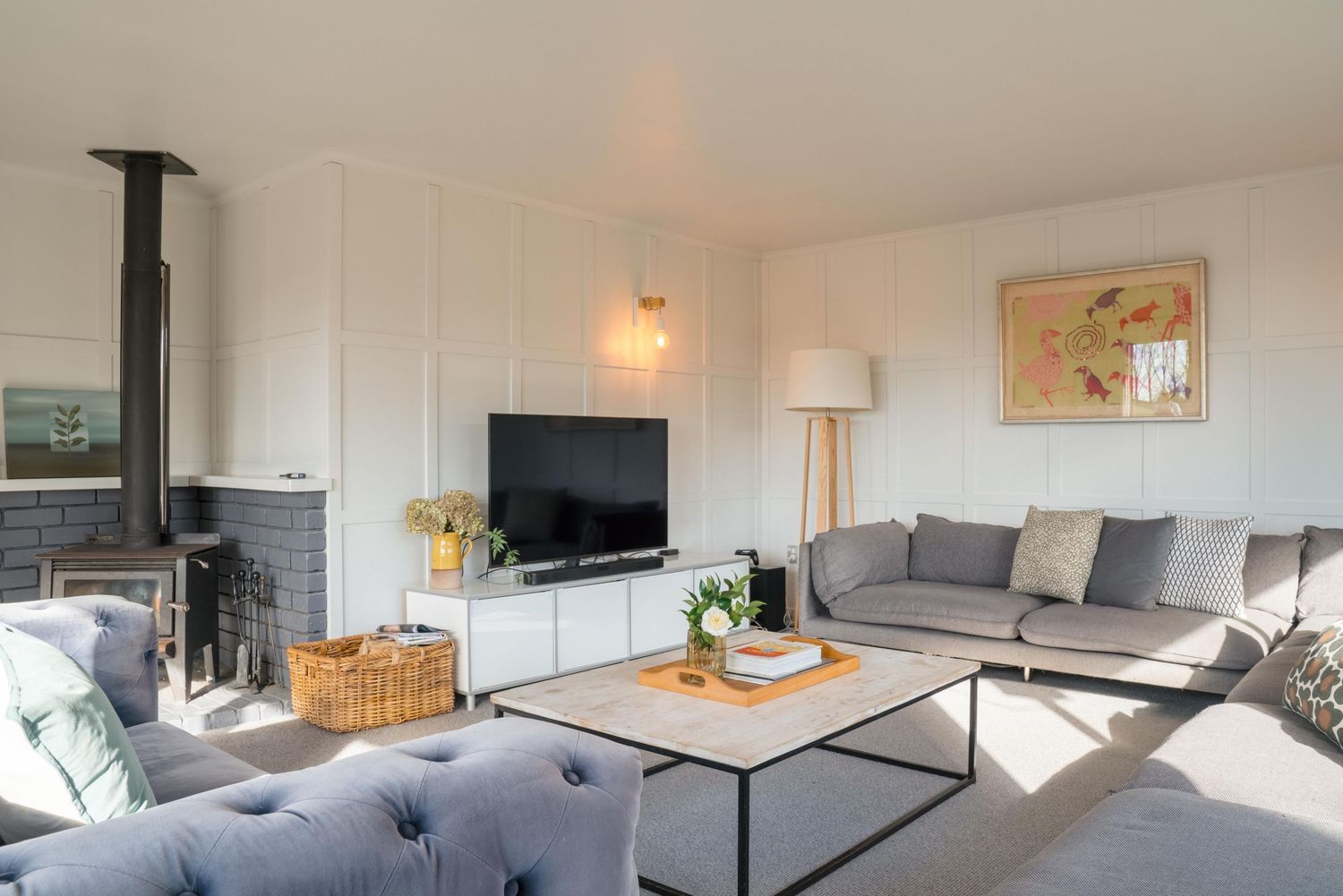 Modern living room interior with gray sofa, wall-mounted TV, wood stove, and natural light streaming through windows.