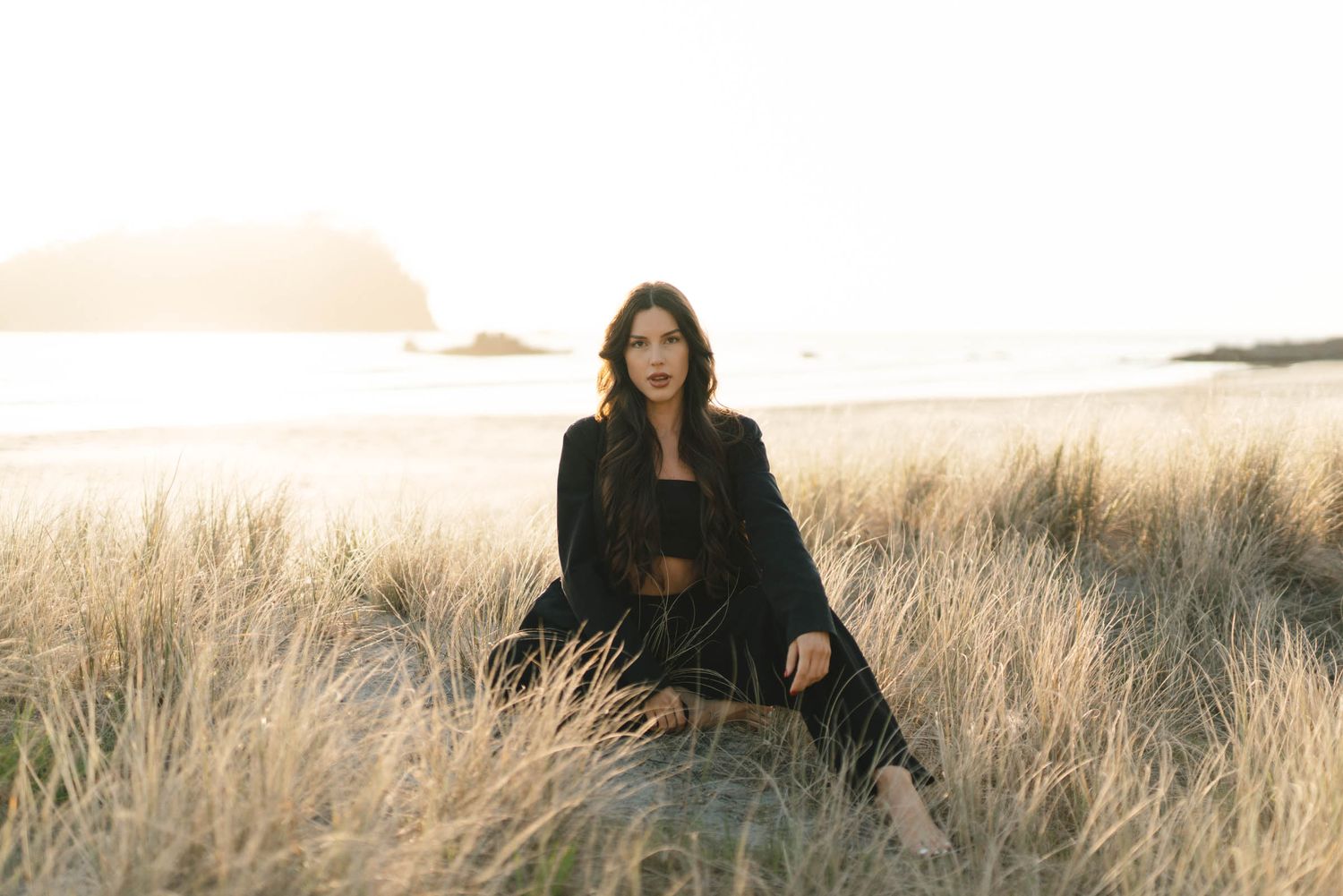 A person in black clothing sits among tall grass with a scenic coastal view in the background.