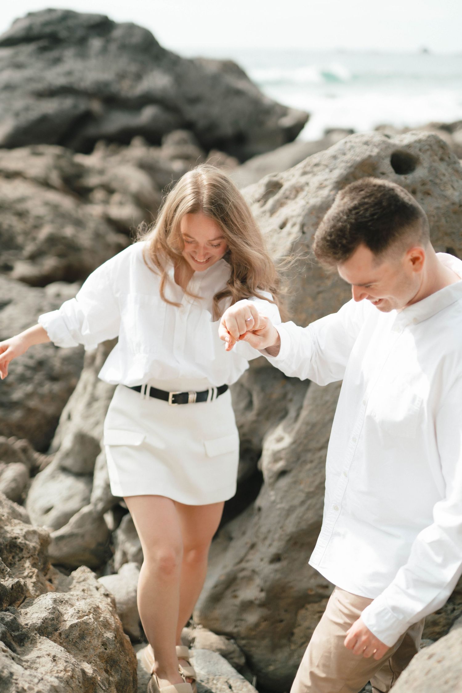 A couple in white outfits playfully posing together on rocky beach terrain.