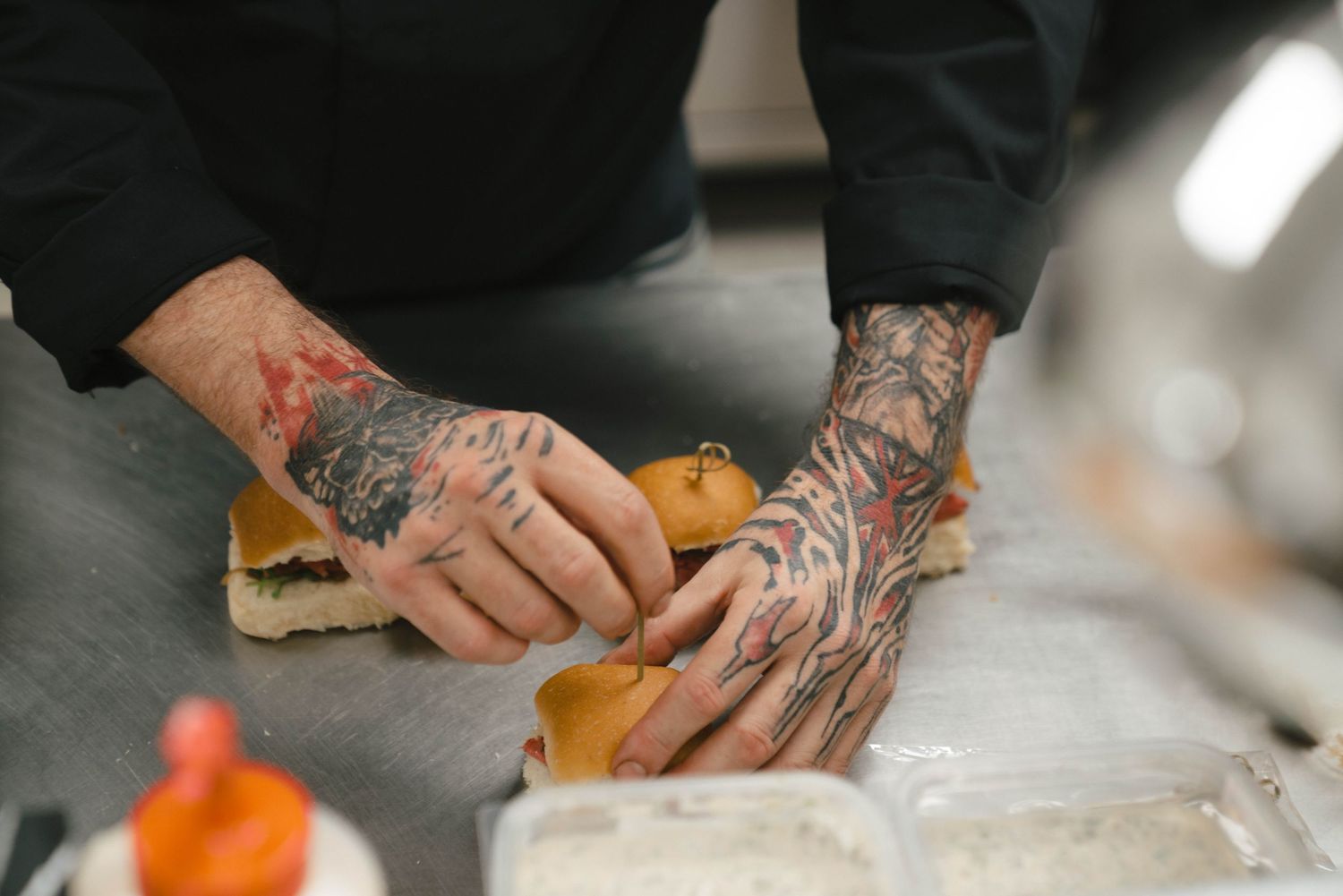 Tattooed hands prepare food on a white countertop with colorful ingredients visible in the background.
