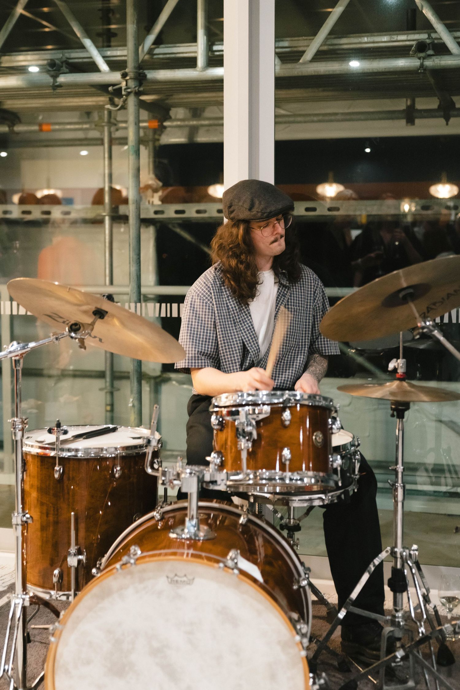 Musician in cap plays drum kit during live performance at indoor venue.