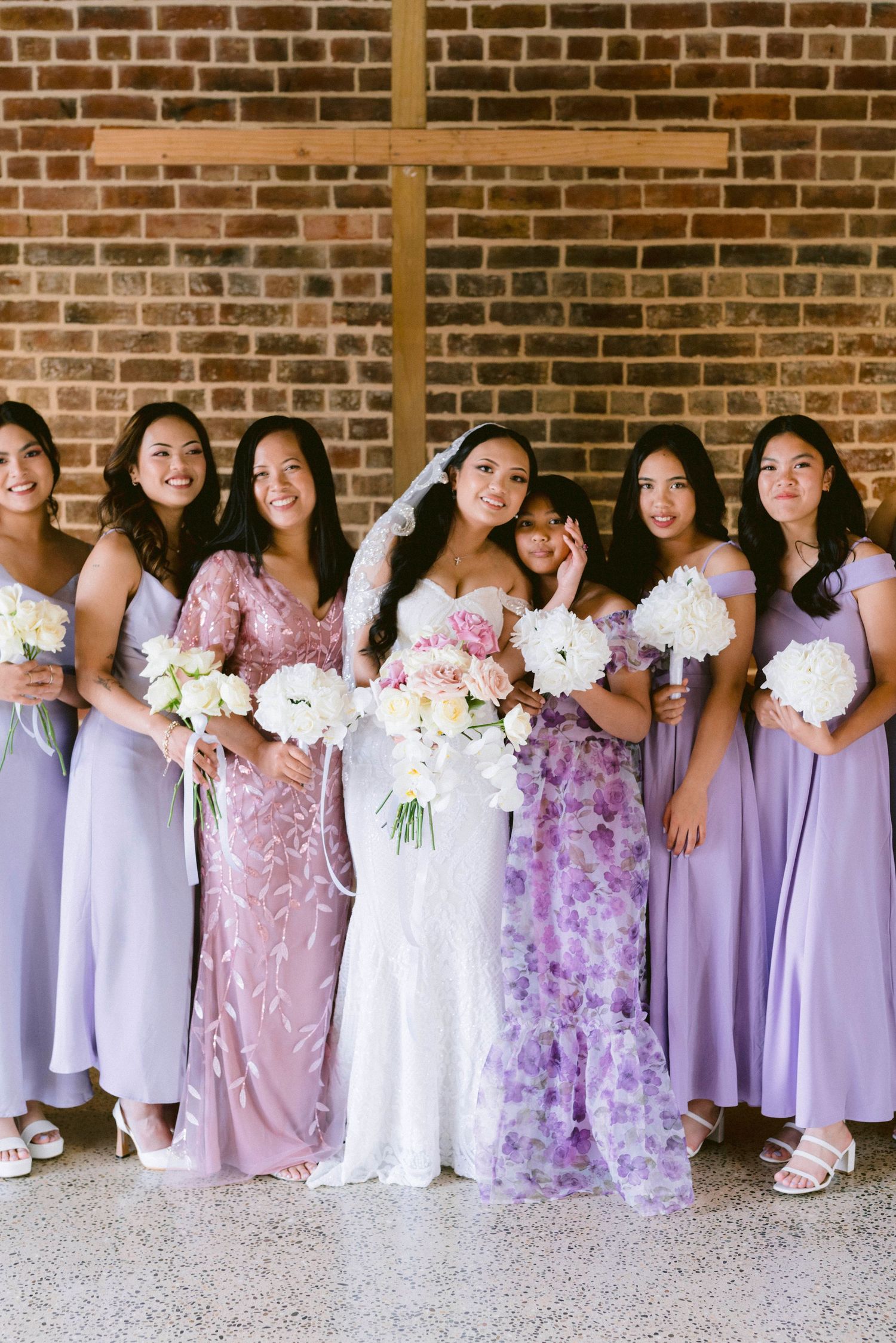 Bridesmaids in lavender and floral dresses pose with white bouquets against brick wall.