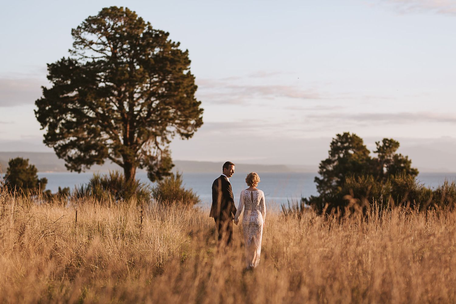 Two people walking through golden prairie grass at sunset with tall trees silhouetted against the sky.