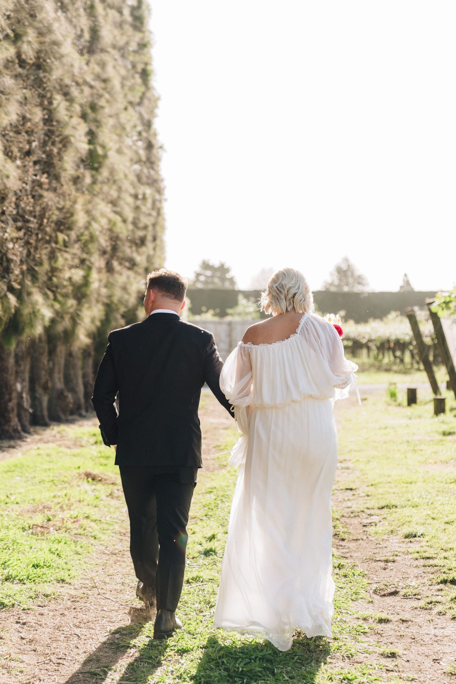 Couple walks away hand in hand down a tree-lined path in formal wedding attire.