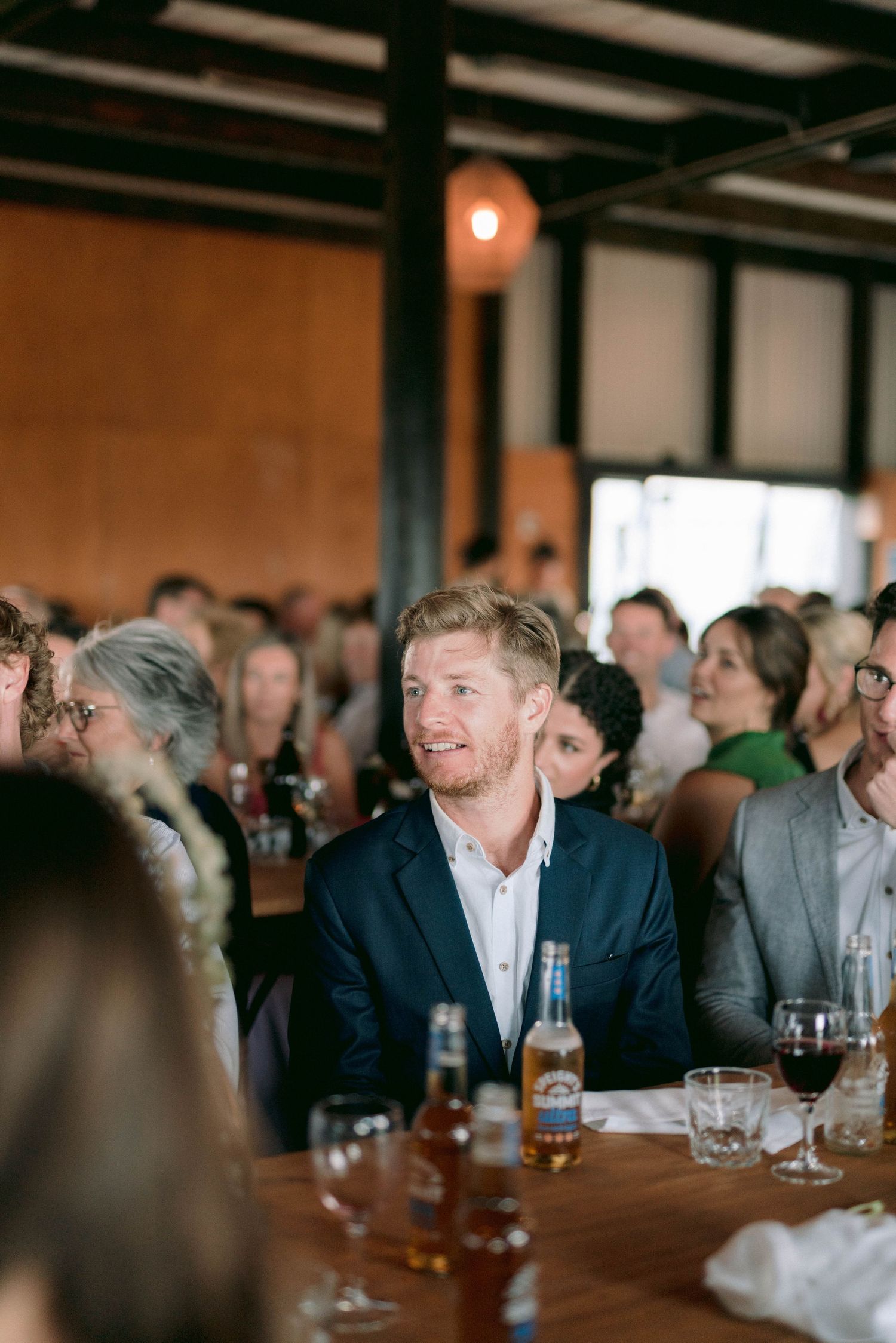 Wedding reception guests gather at wooden tables in a rustic indoor venue with exposed beams.