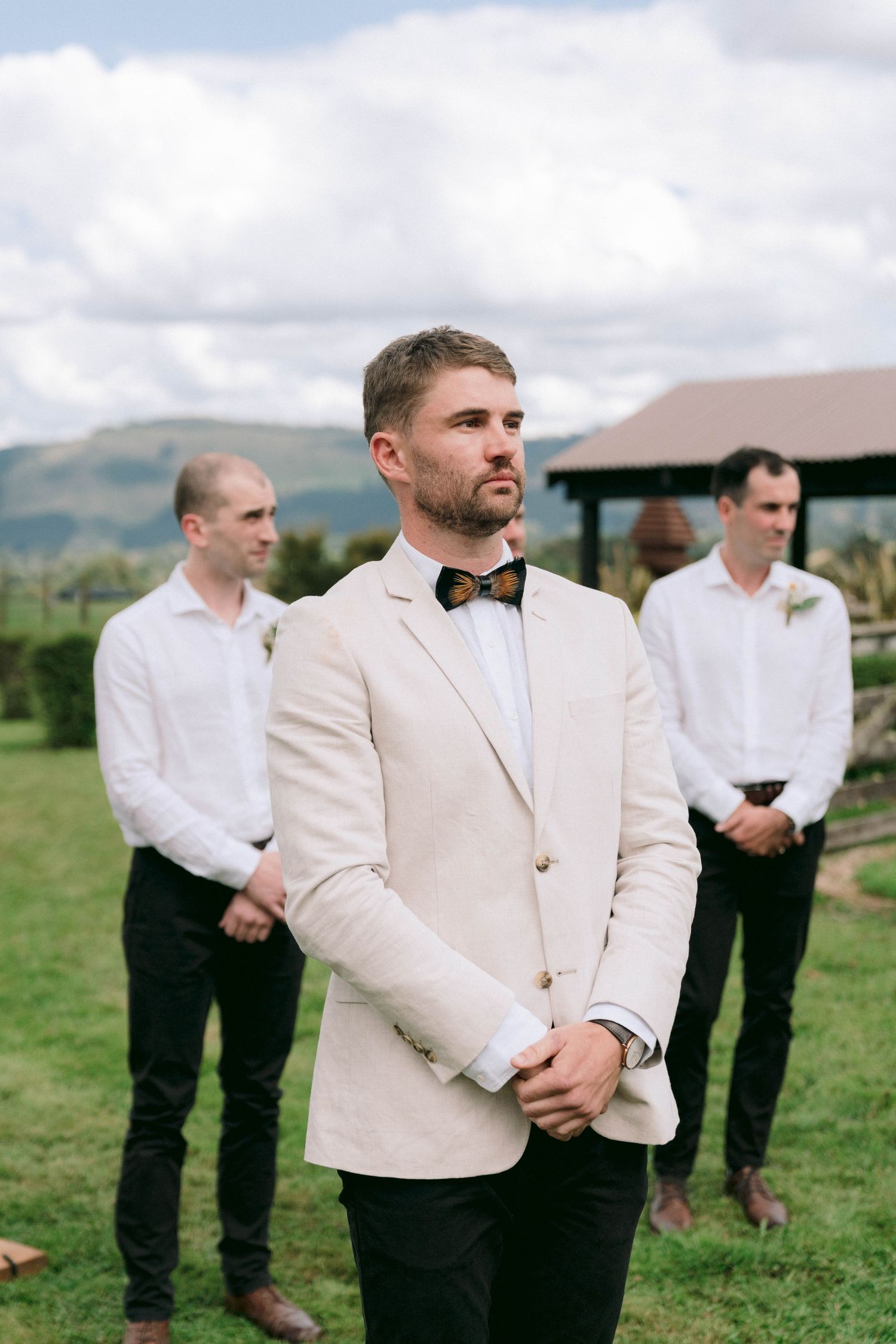 Formal wedding party members stand on grass with mountains in background as groomsman poses in beige jacket.