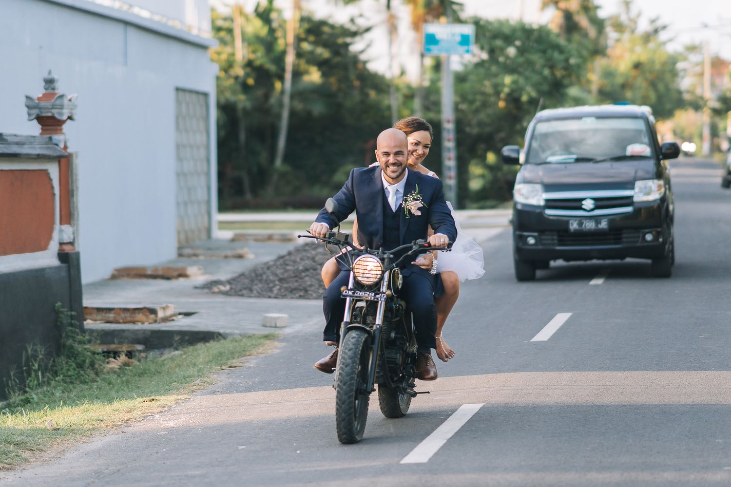 Two people enjoying a motorcycle ride together on a sunny street with palm trees in the background.