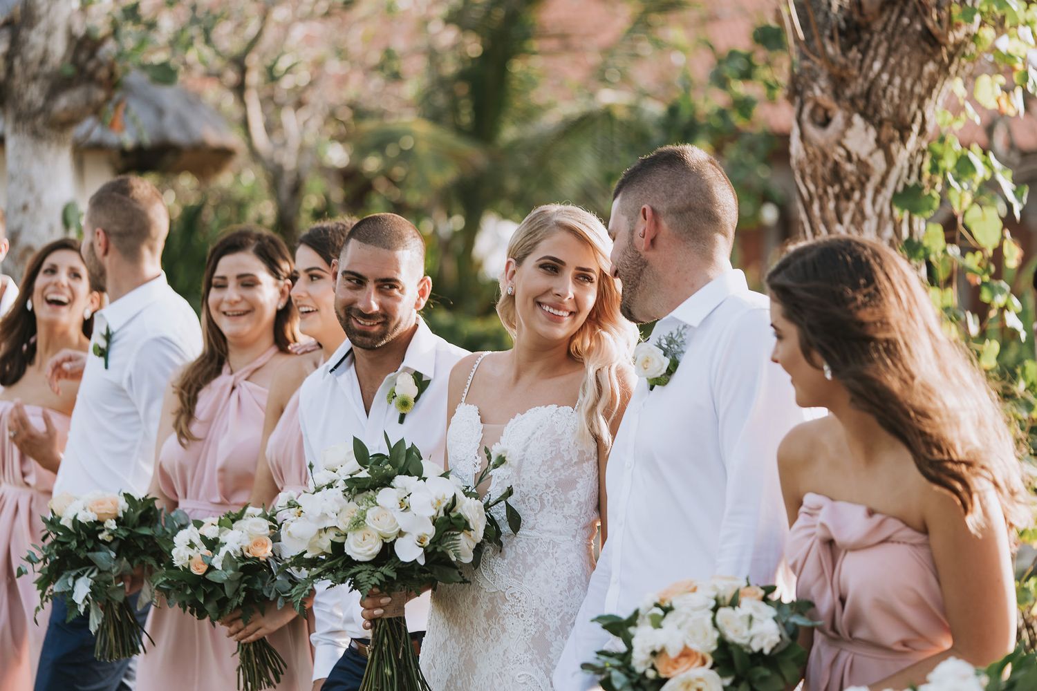 Wedding party in white and blush pink attire posing with floral bouquets outdoors.